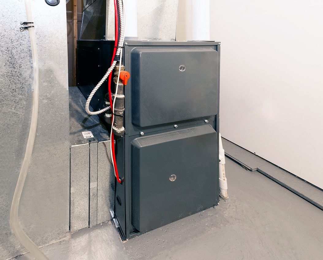 A gray HVAC furnace unit sits on a concrete floor next to a silver metal duct in a room with white walls.