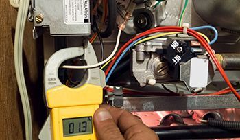 Hand using a clamp meter to measure the current on a gas furnace's wire. The display reads 0.3, showing electrical usage.