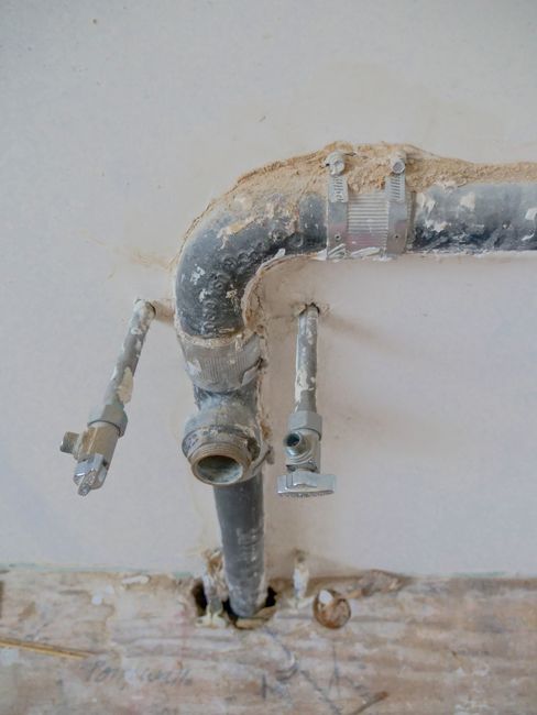 Corroded metal pipe with two open side valves against a wall above a wooden floor