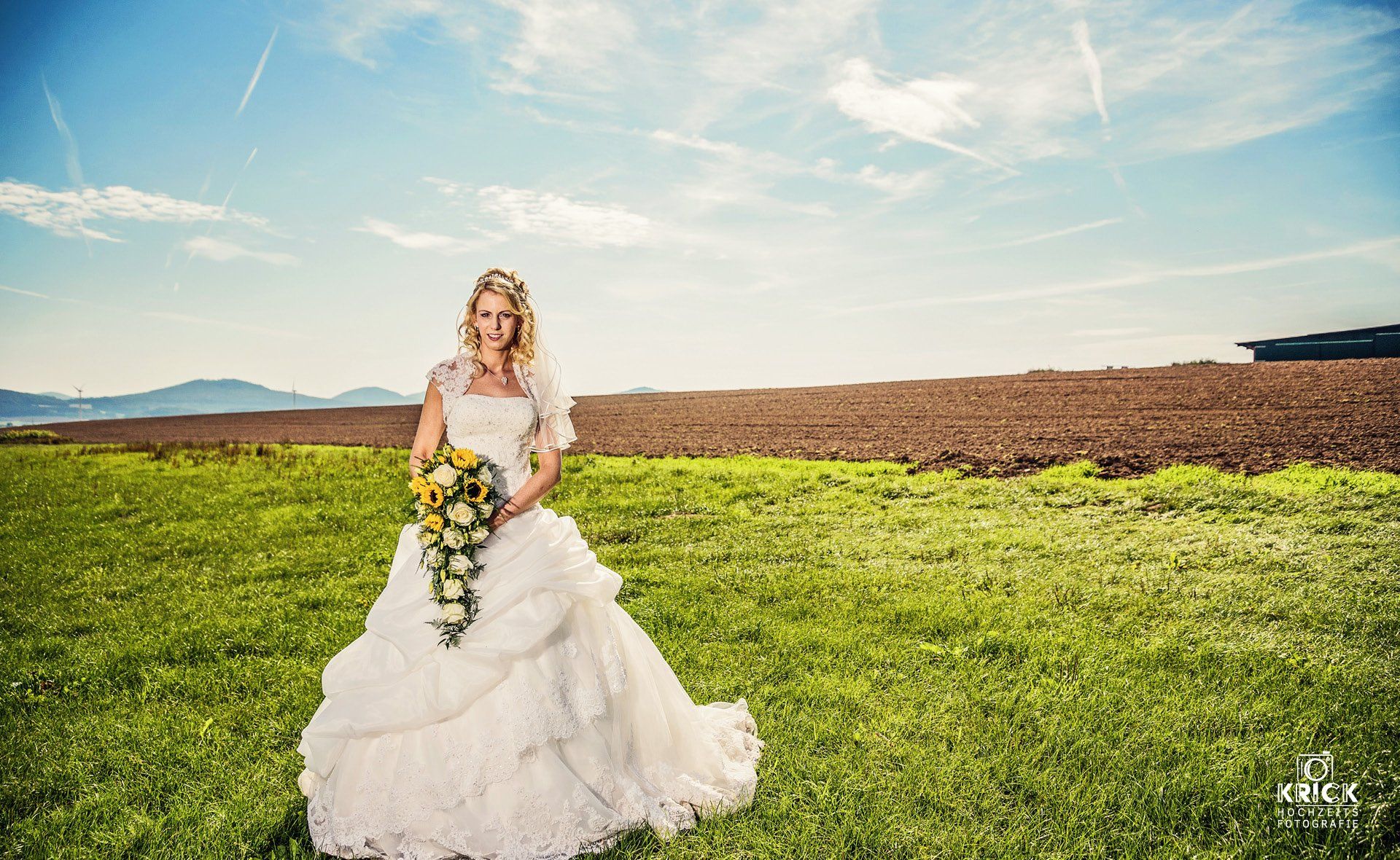 Eine Braut in einem weißen Kleid hält einen gelben Brautstrauß auf einer grasbewachsenen Wiese unter strahlend blauem Himmel.