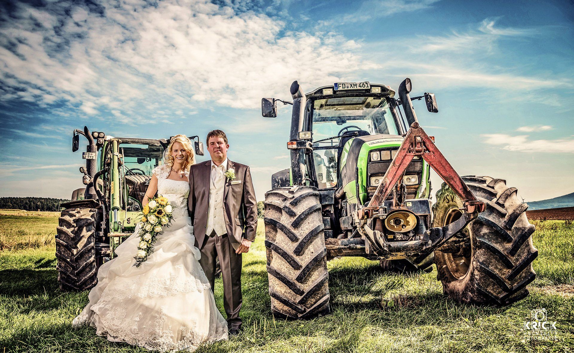 Ein Hochzeitspaar steht zwischen zwei Traktoren auf einem Feld unter blauem Himmel