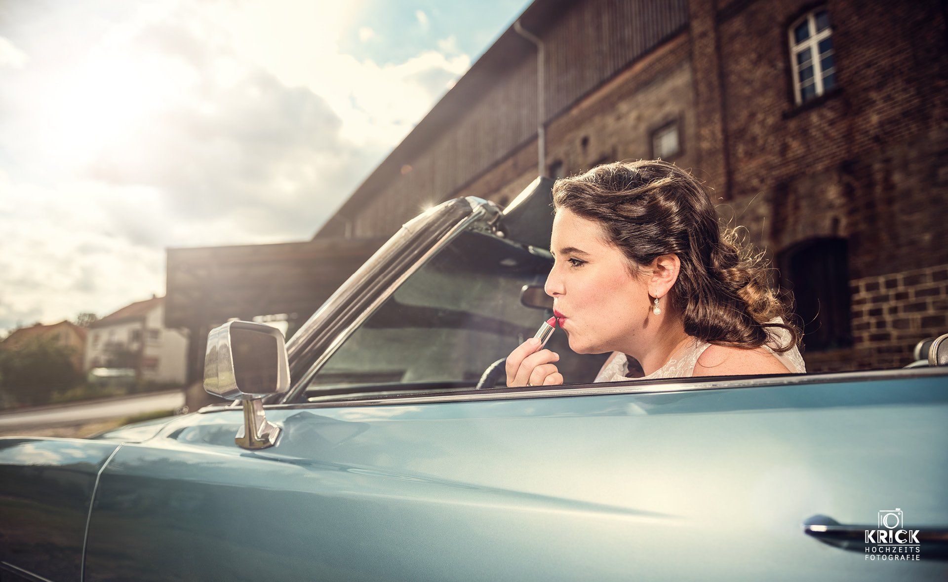 Eine Frau fährt in einem blauen Oldtimer-Cabriolet auf einer sonnenbeschienenen Straße; im Hintergrund ist ein Backsteingebäude zu sehen.