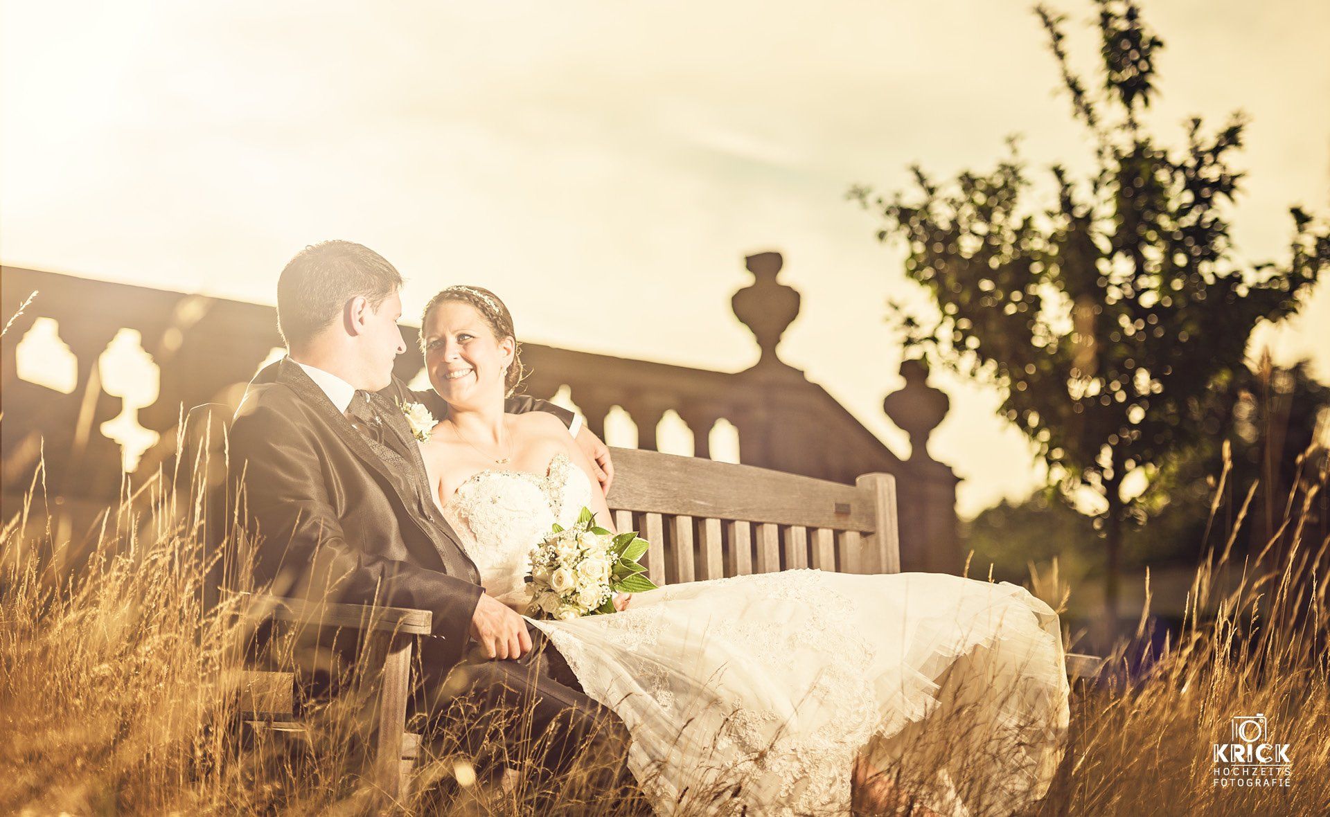 Ein Paar in Hochzeitskleidung sitzt bei Sonnenuntergang im Freien auf einer rustikalen Bank und hält einen Brautstrauß.