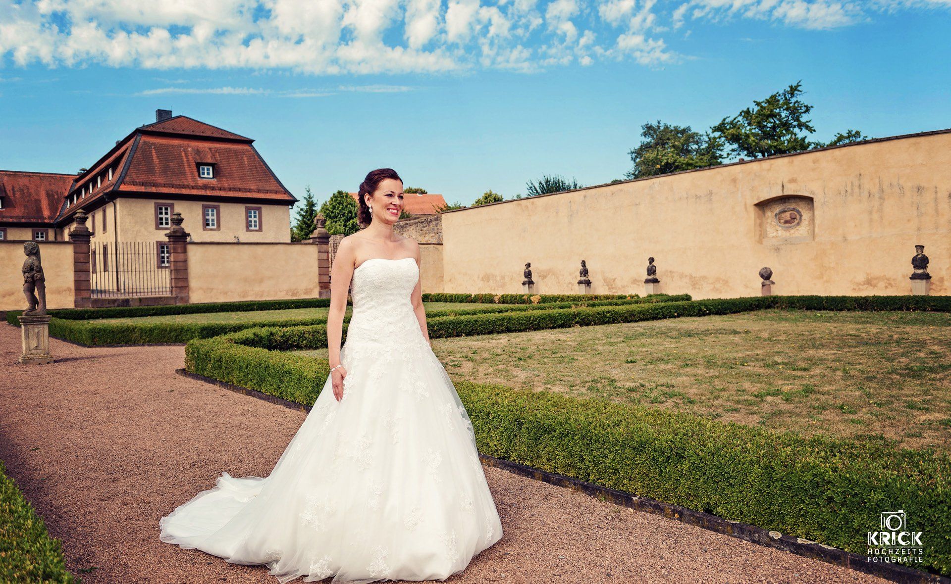 Eine Braut in einem weißen Hochzeitskleid steht auf einem Weg neben einem historischen Gebäude und einer Steinmauer unter blauem Himmel.