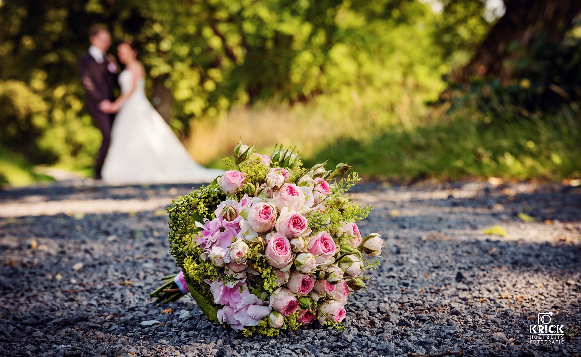 Ein Strauß rosa und weißer Blumen auf Kies, im Hintergrund ein verschwommenes Brautpaar – im Freien