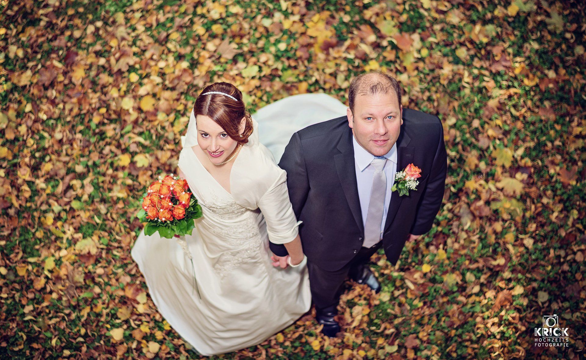 Braut und Bräutigam halten Händchen und blicken vor einem herbstlich belaubten Bodendecker nach oben.