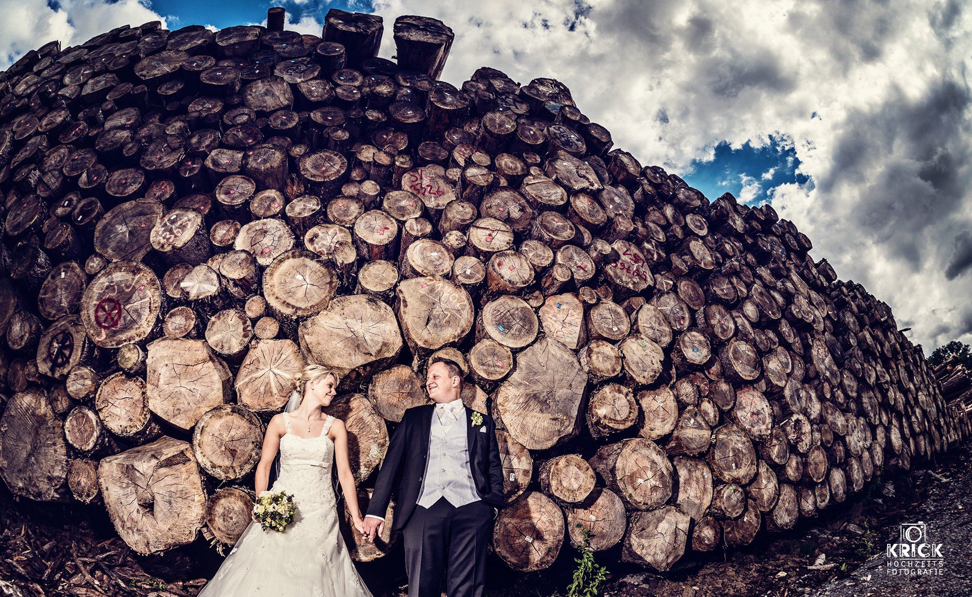 Frischvermählte in Hochzeitskleidung stehen vor einem riesigen Holzstapel unter einem bewölkten Himmel