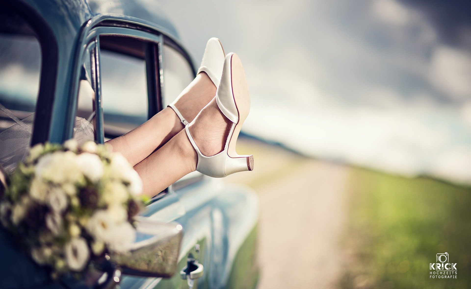 Die Füße der Braut in weißen High Heels ruhen auf einer Autotür, davor ein Brautstrauß und die verschwommene Straße.