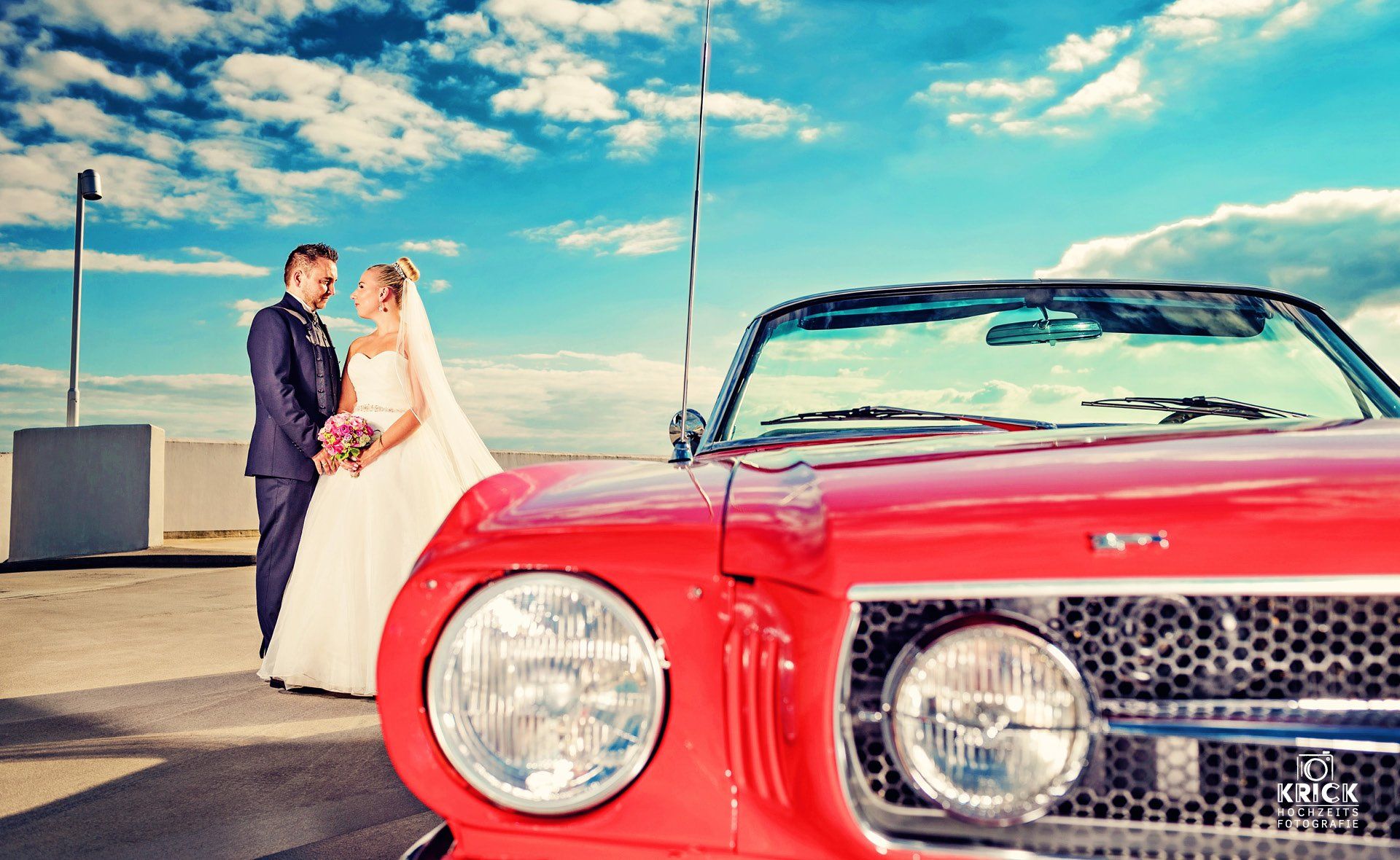 Braut und Bräutigam stehen neben einem roten Oldtimer unter blauem Himmel