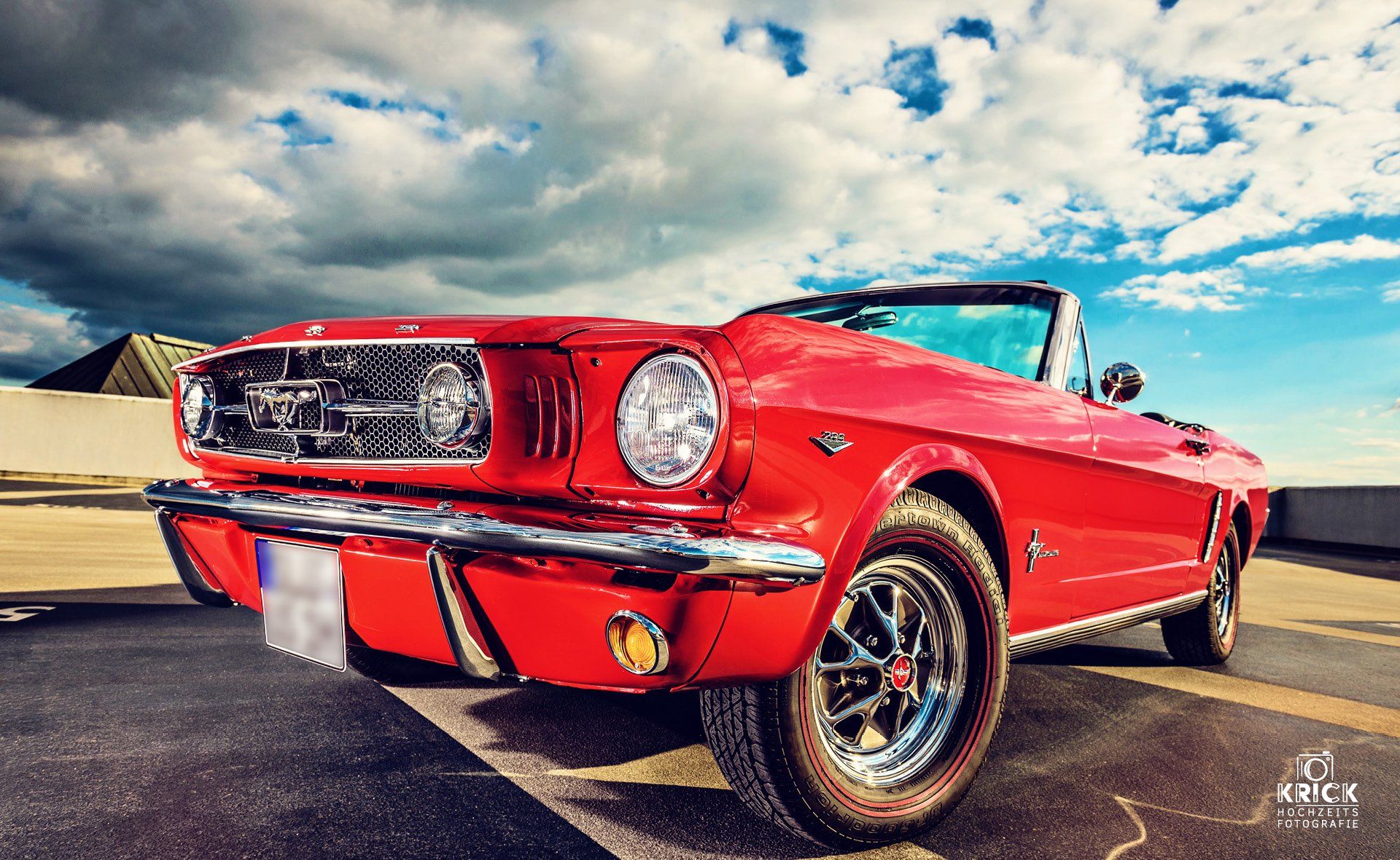 Ein rotes, klassisches Mustang-Cabriolet parkt auf einer Straße unter einem bewölkten blauen Himmel.