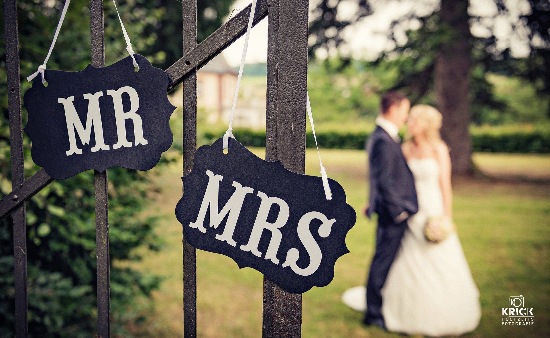 An einem Zaun hängen Schilder mit der Aufschrift „MR“ und „MRS“, im Hintergrund ist ein Brautpaar verschwommen zu sehen.