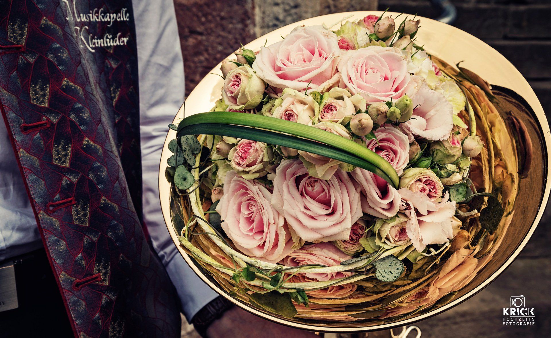 Ein Strauß aus rosa und weißen Rosen mit Grünzeug in einem goldenen Korb, neben einem dunkel gemusterten Stoff.