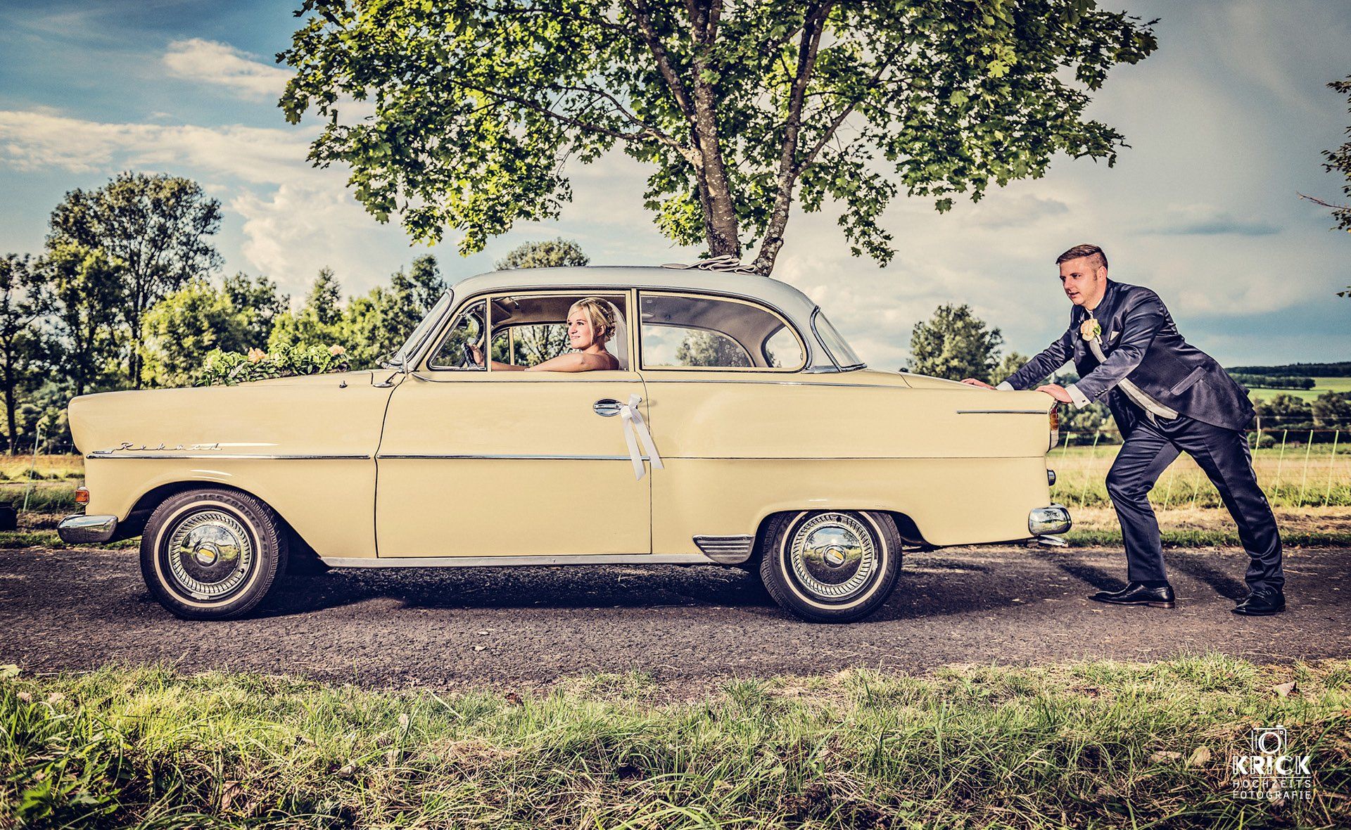 Ein cremefarbener Oldtimer steht auf einer Wiese unter einem belaubten Baum. Ein Mann schiebt ihn, und eine Person sitzt darin.
