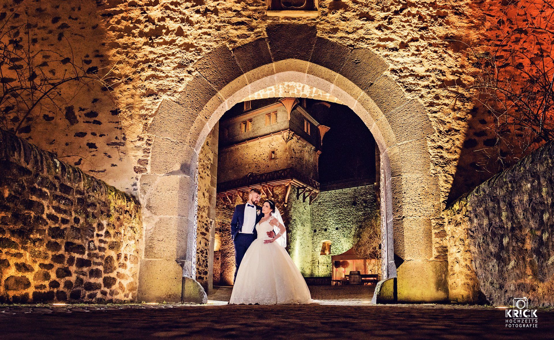 Braut und Bräutigam unter einem Steinbogen in einem warm beleuchteten historischen Innenhof bei Nacht