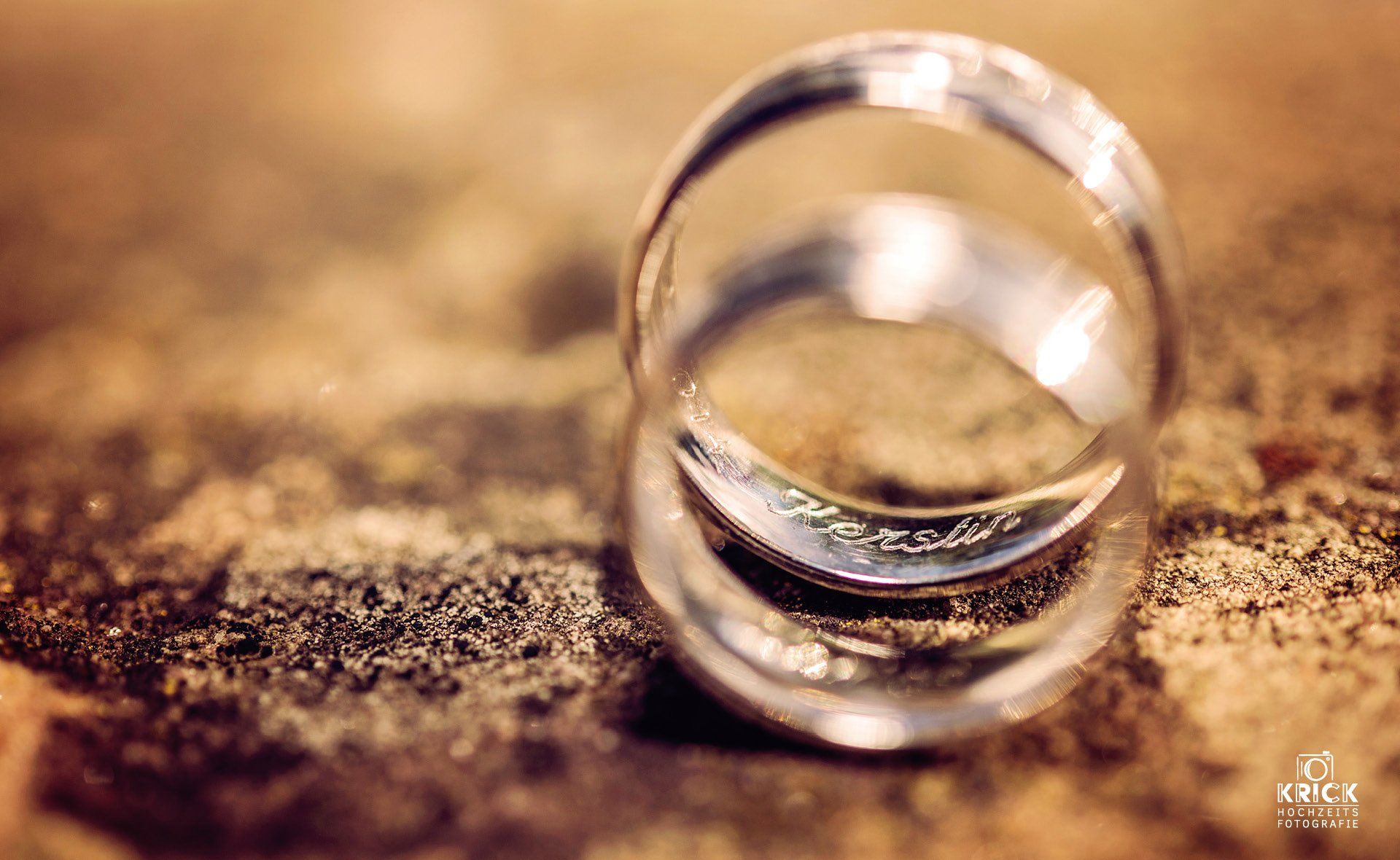 Zwei goldene Ringe stehen aufrecht auf einer strukturierten braunen Oberfläche vor einem warmen, verschwommenen Hintergrund.