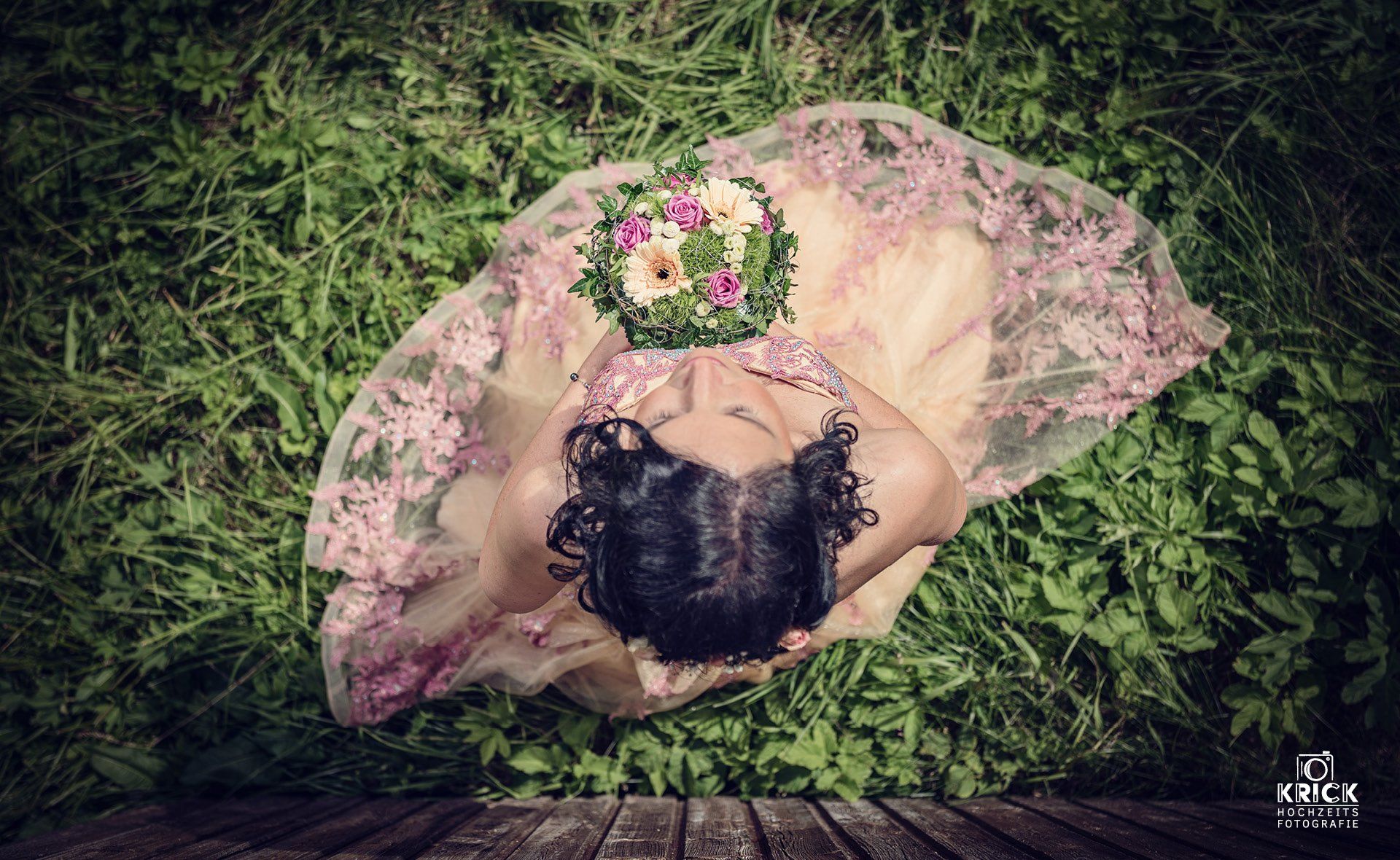 Frau in einem rosa Kleid, die einen Blumenkranz hält, von oben vor einem grasbewachsenen Hintergrund gesehen.