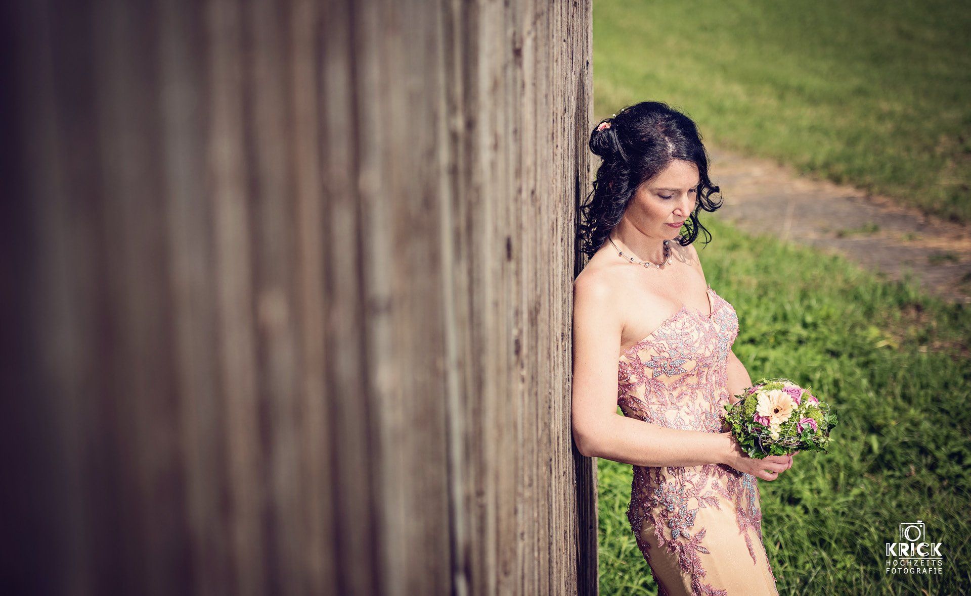 Eine Frau in einem rosa geblümten Kleid lehnt im Freien an einer Holzwand und hält einen kleinen Blumenstrauß.