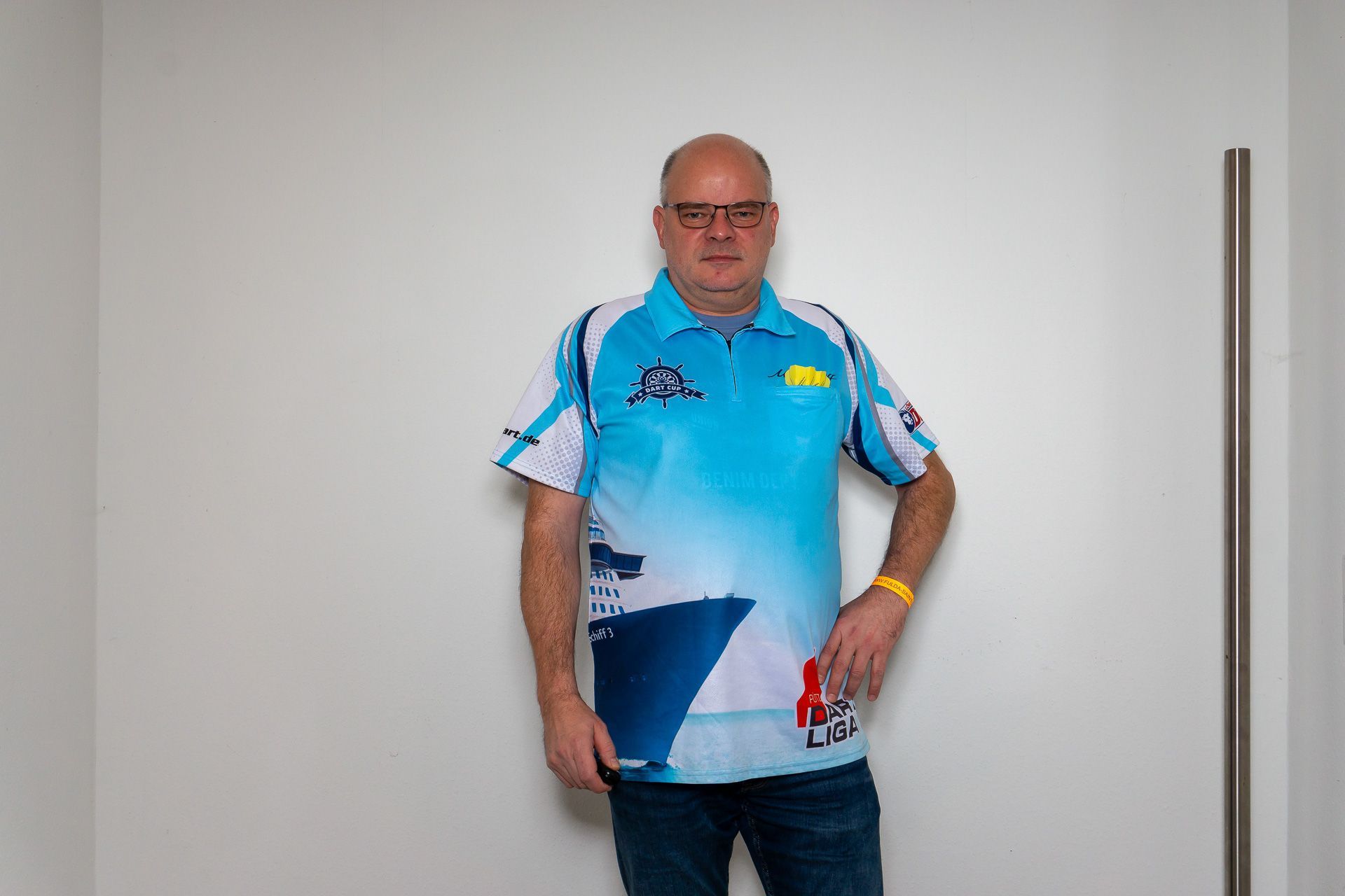 Ein Mann, der an einer schlichten Wand steht, trägt ein blaues Sporthemd und Jeans.