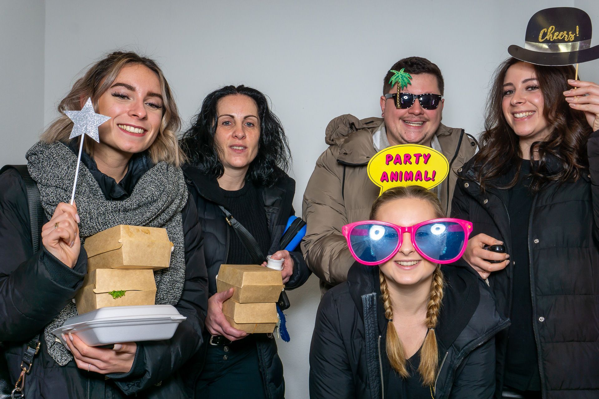 Fünf Personen posieren drinnen, lächeln und halten Requisiten, darunter einen gelben „PARTY ANIMAL“-Hut und eine pinkfarbene Sonnenbrille.