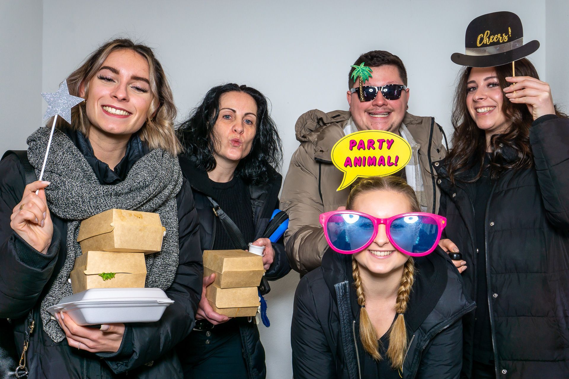 Fünf Personen posieren mit gestapelten Kisten, eine trägt eine übergroße pinkfarbene Sonnenbrille und eine andere einen ausgefallenen Hut in einem Studio.