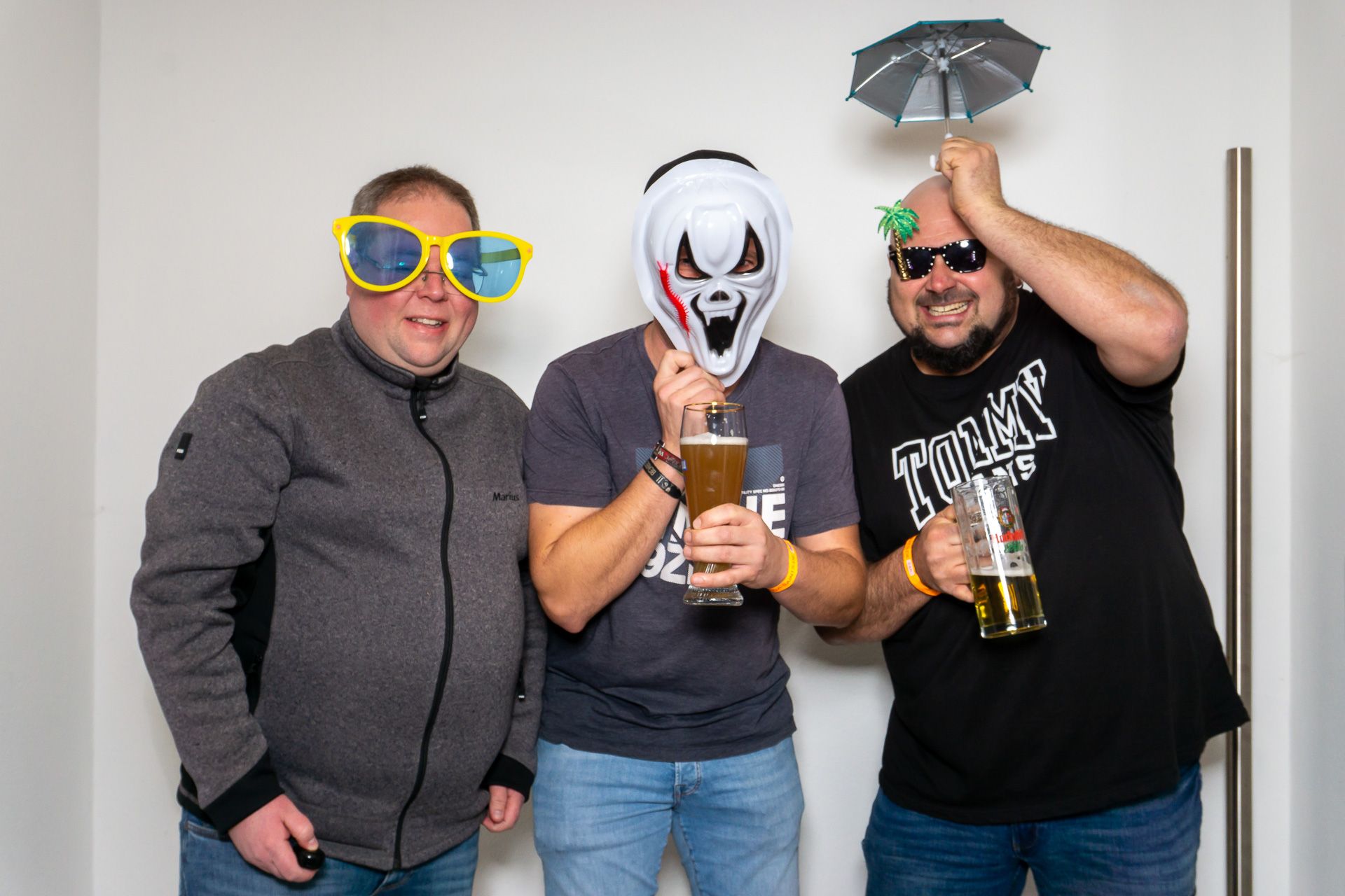 Drei Personen posieren drinnen mit Halloween-Requisiten, tragen alberne Brillen und Masken und halten Getränke in der Hand.