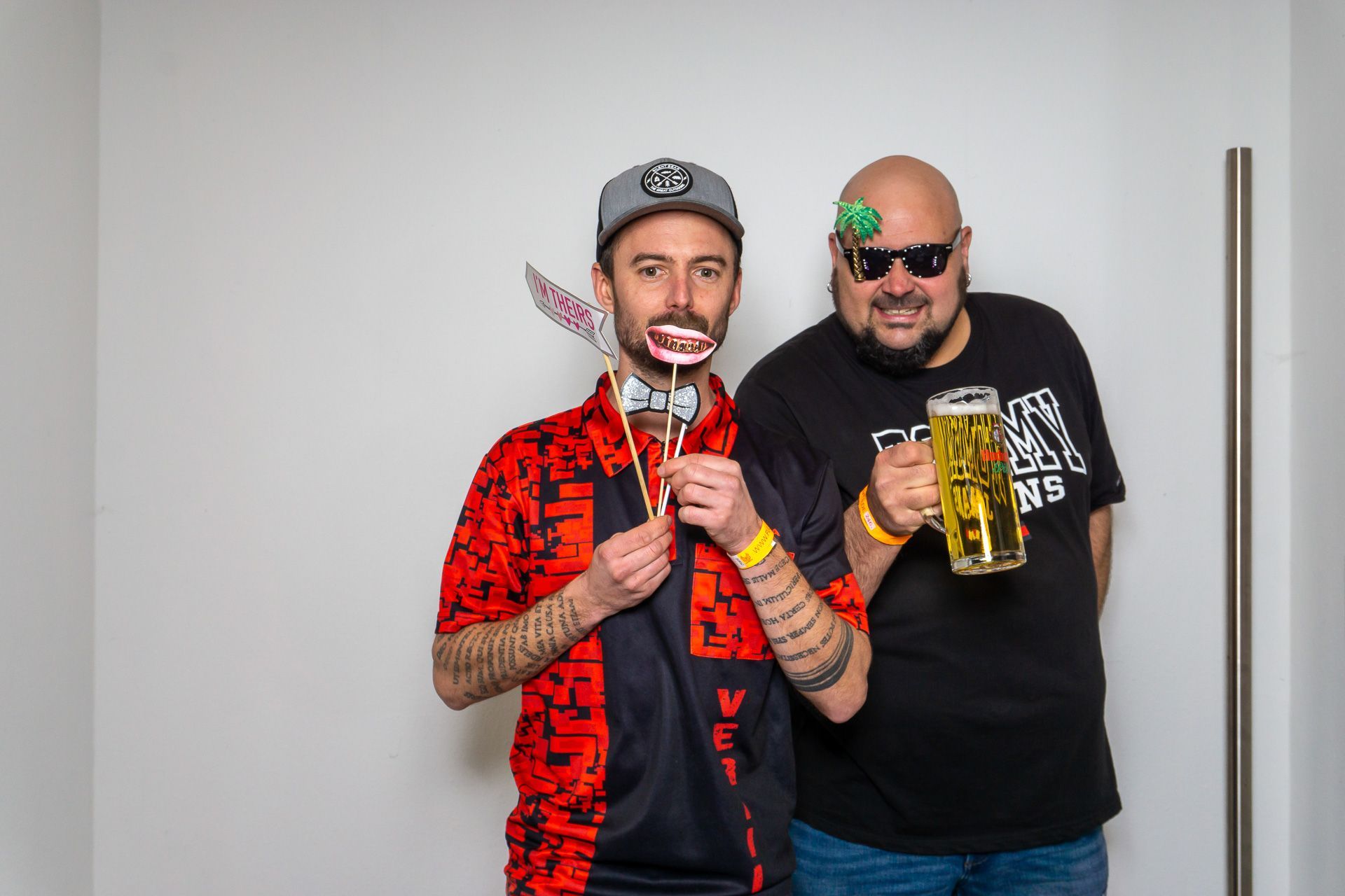 Zwei lächelnde Männer posieren vor einer schlichten Wand, einer hält einen Lutscher, der andere einen Becher und eine Dose.