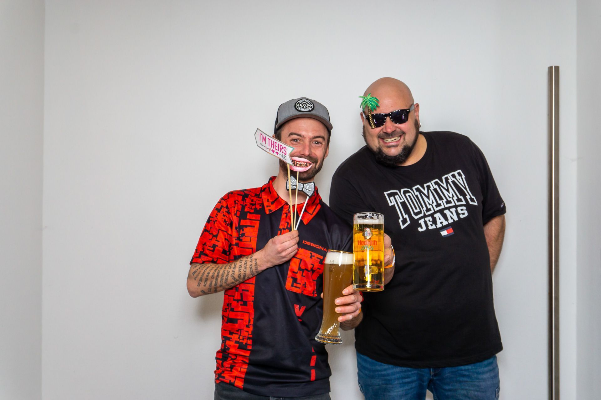 Zwei lächelnde Männer mit Bierkrügen in der Hand stehen vor einer weißen Wand.