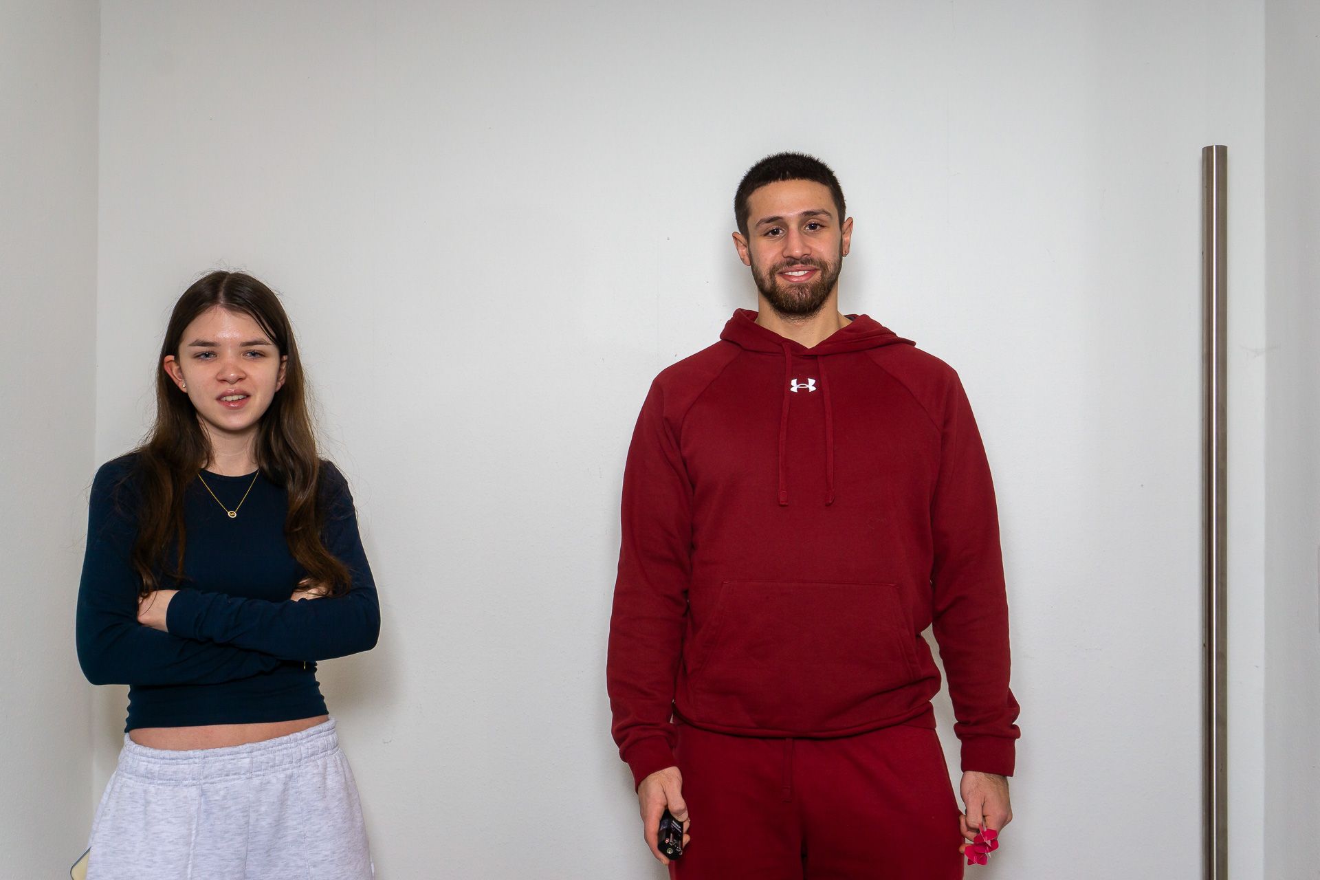 Zwei Personen stehen vor einer schlichten Wand, eine trägt ein dunkelblaues Oberteil und eine weiße Hose, die andere einen roten Kapuzenpullover.