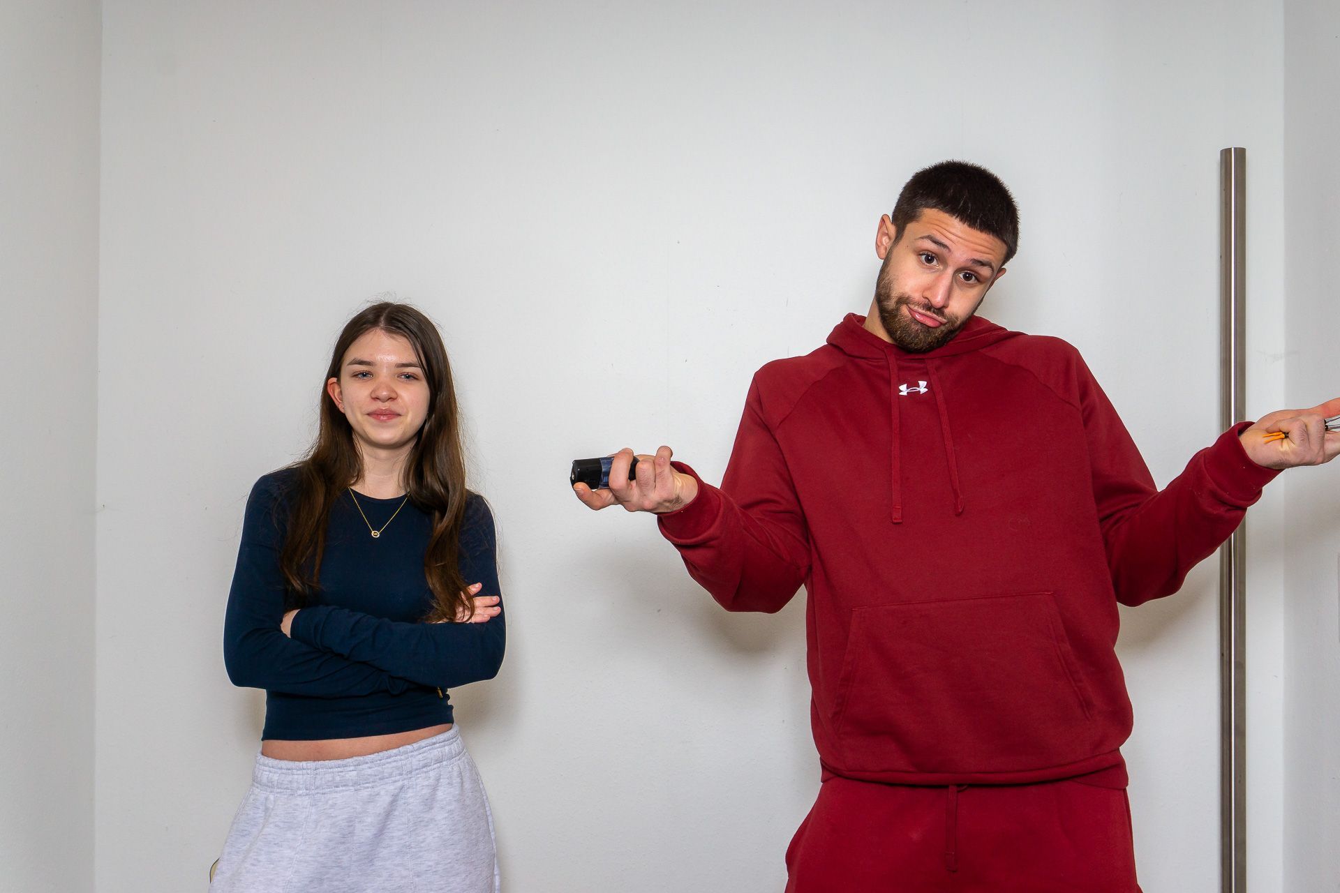 Zwei Personen stehen an einer schlichten Wand, eine mit verschränkten Armen, die andere in rotem Trainingsanzug, die Achseln zuckend.