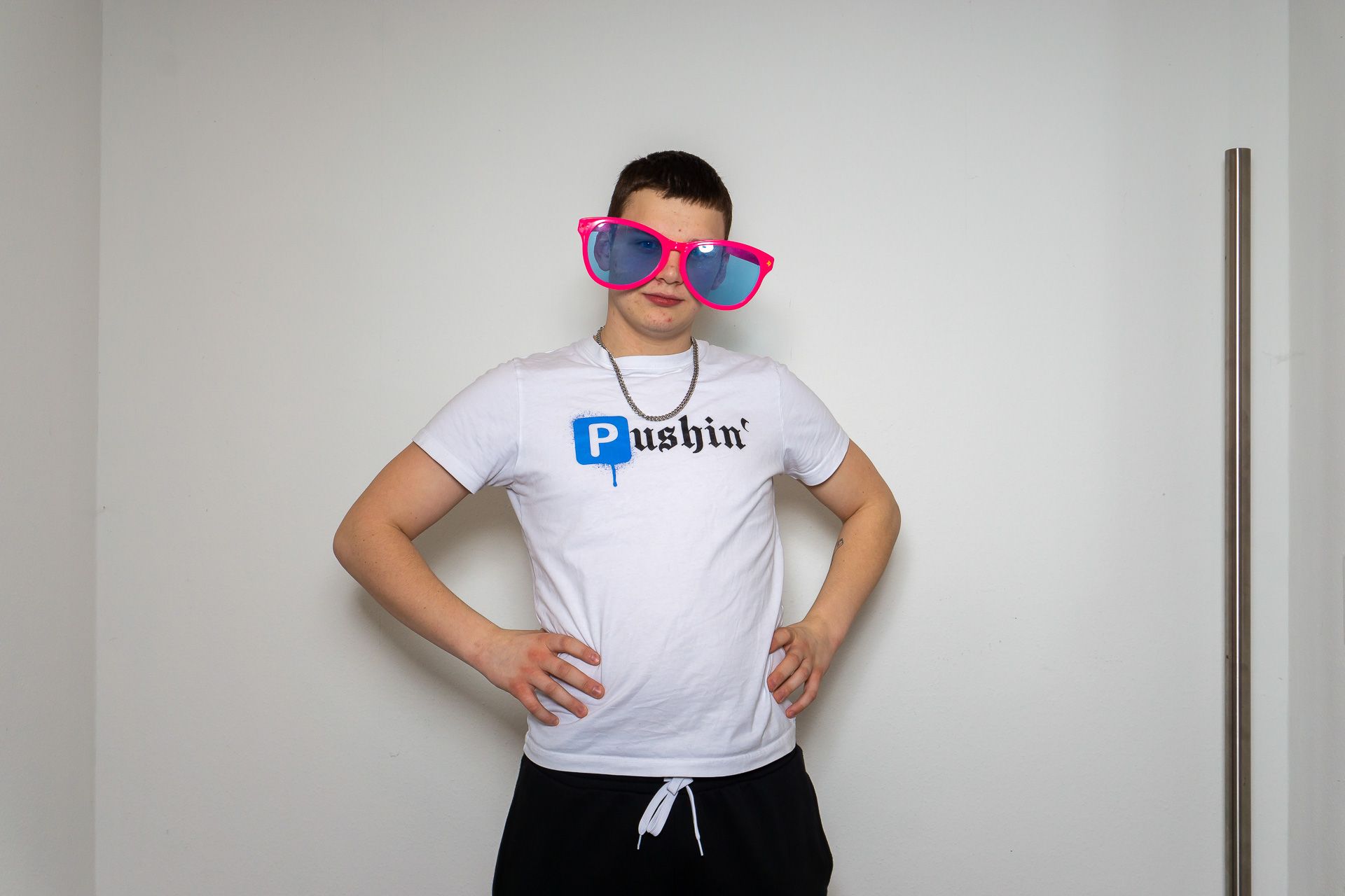 Eine Person posiert in einem Innenraum mit den Händen in den Hüften. Sie trägt ein weißes T-Shirt, eine schwarze Hose und eine leuchtend pinkfarbene Sonnenbrille.