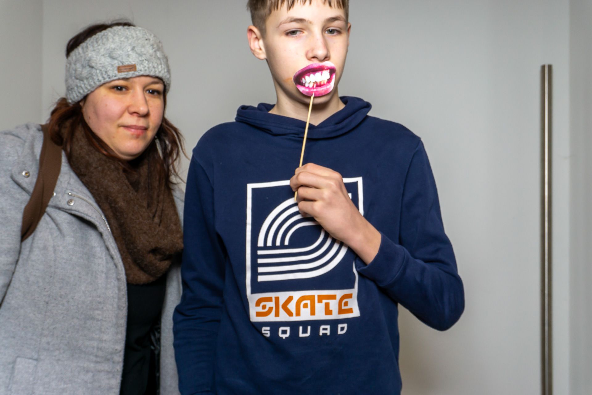 Zwei Personen stehen in einem Innenraum; ein Junge in einem dunkelblauen Kapuzenpulli hält einen rosa Lutscher an seinen Mund.