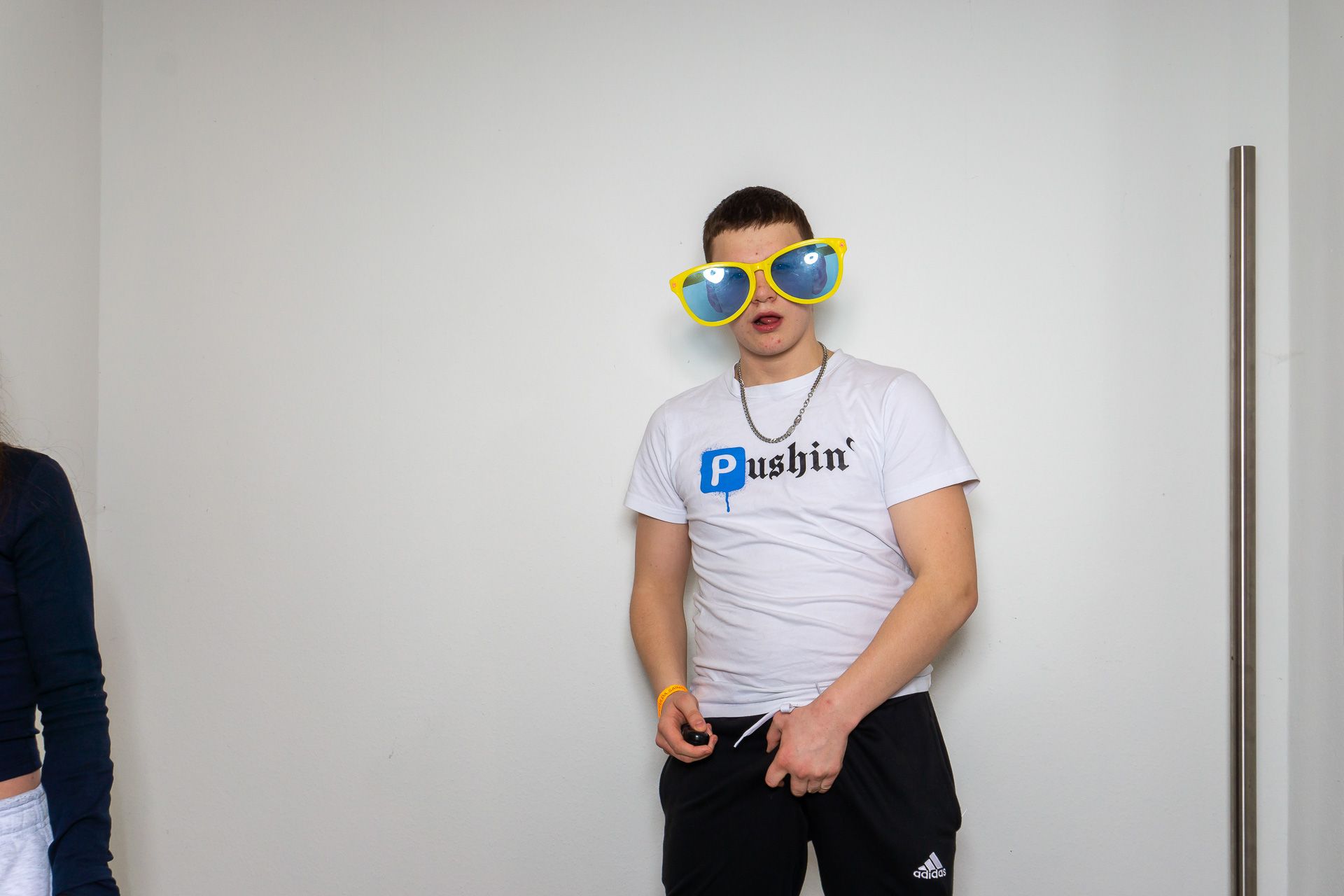 Eine Person in einem weißen T-Shirt und einer übergroßen, auffälligen Sonnenbrille steht vor einer leeren Wand.
