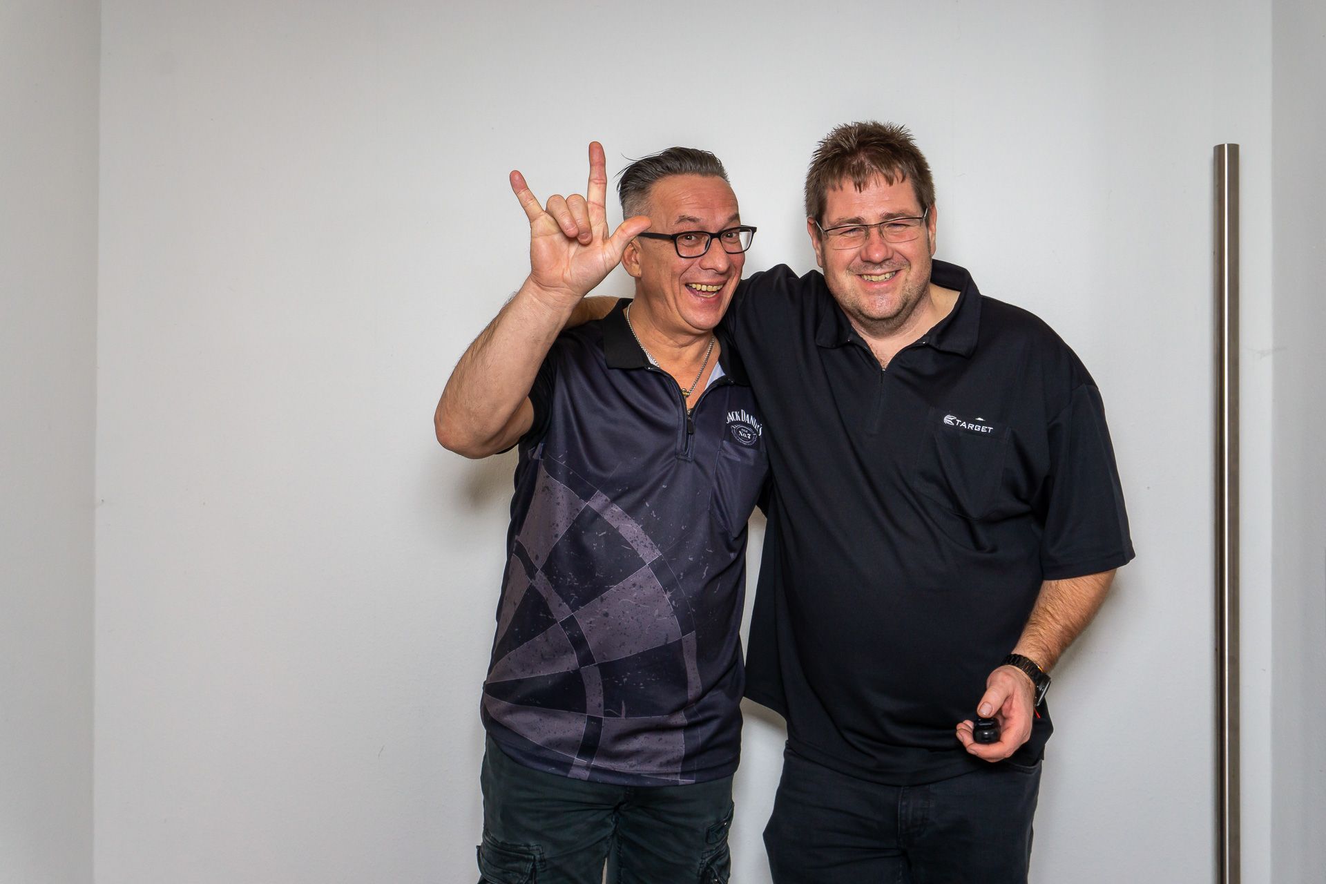 Zwei Männer stehen lächelnd nebeneinander vor einer schlichten weißen Wand, einer von ihnen macht ein Handzeichen.