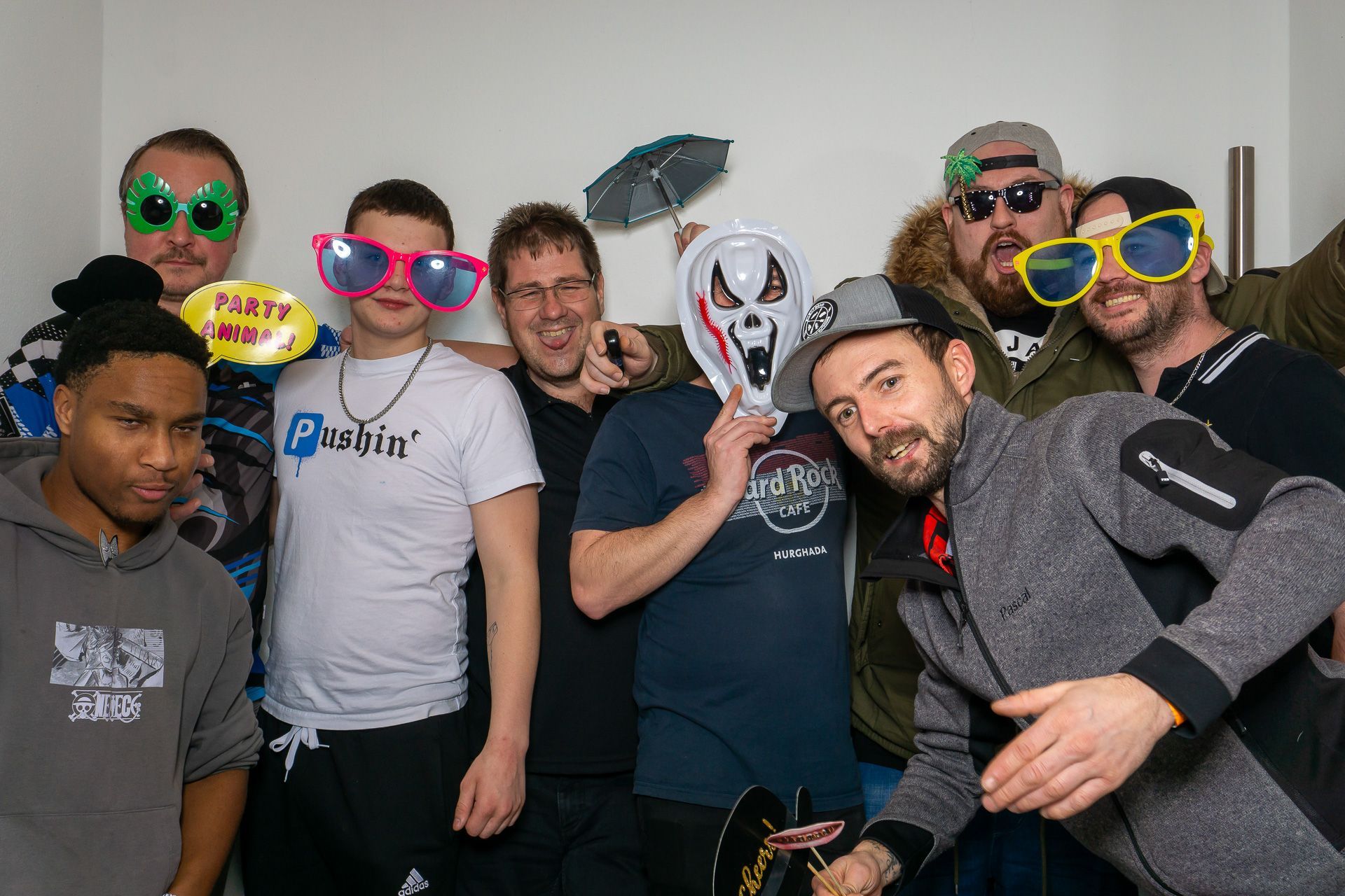 Eine Gruppe von Menschen posiert in einem Innenraum; einige tragen bunte, ausgefallene Sonnenbrillen, einer eine weiße Maske.