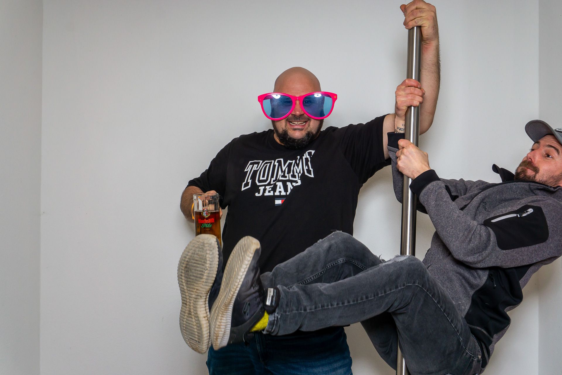 Zwei Männer lachen und posieren mit übergroßen pink-blauen Sonnenbrillen, einer hält drinnen eine Stange.