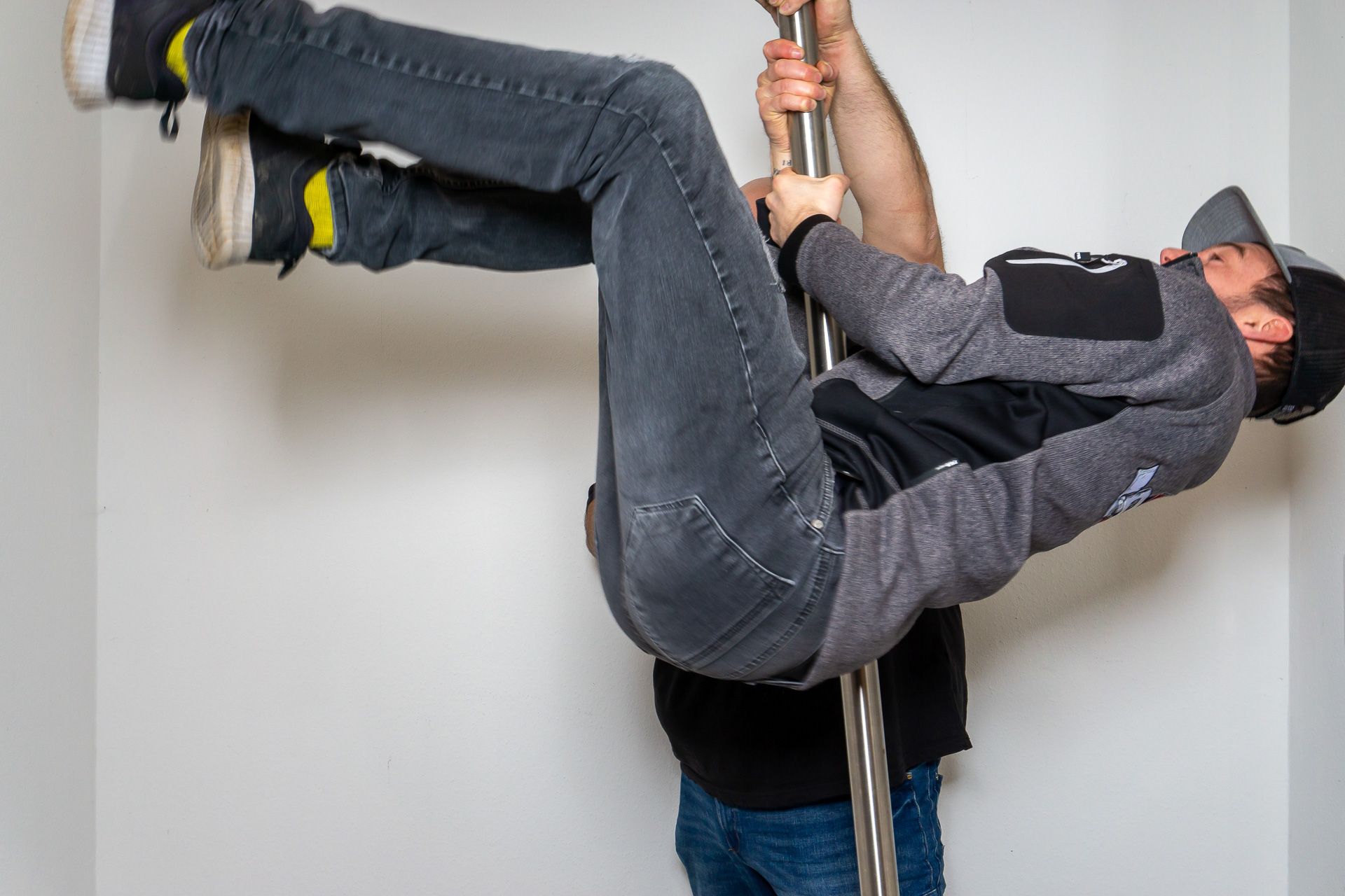 Eine Person, die Jeans und eine Kappe trägt, hängt waagerecht an einer Stange in einem Raum und wird von einer anderen Person darunter gehalten.