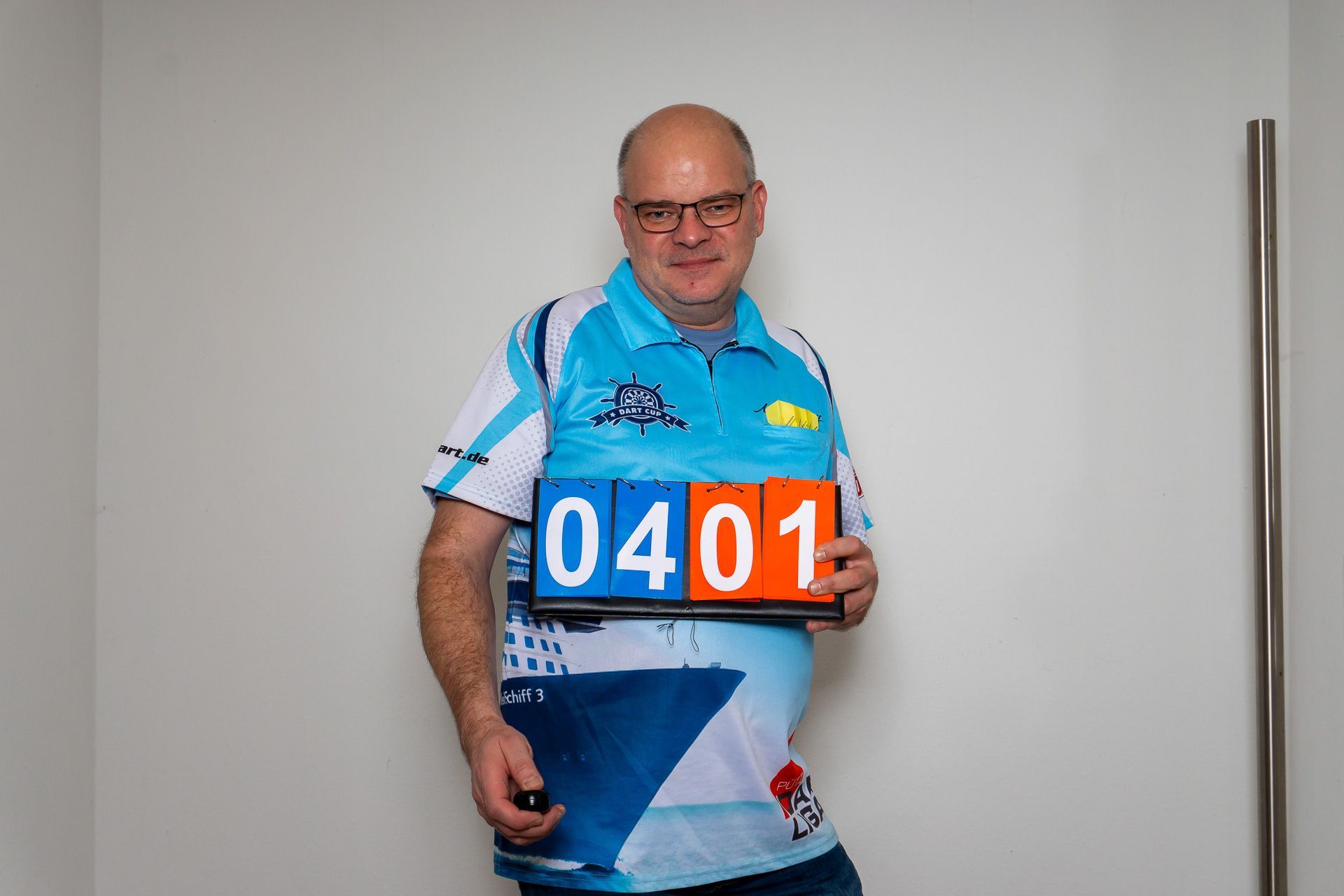 Ein Mann hält ein blau-oranges Startnummernschild und steht in einem Innenraum vor einer schlichten Wand.