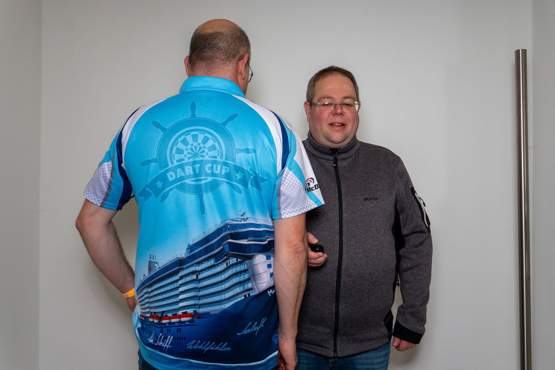 Zwei Männer stehen in einem Innenraum, einer in einem blauen Hemd, von hinten zu sehen, der andere in einer grauen Jacke, nach vorne gewandt.
