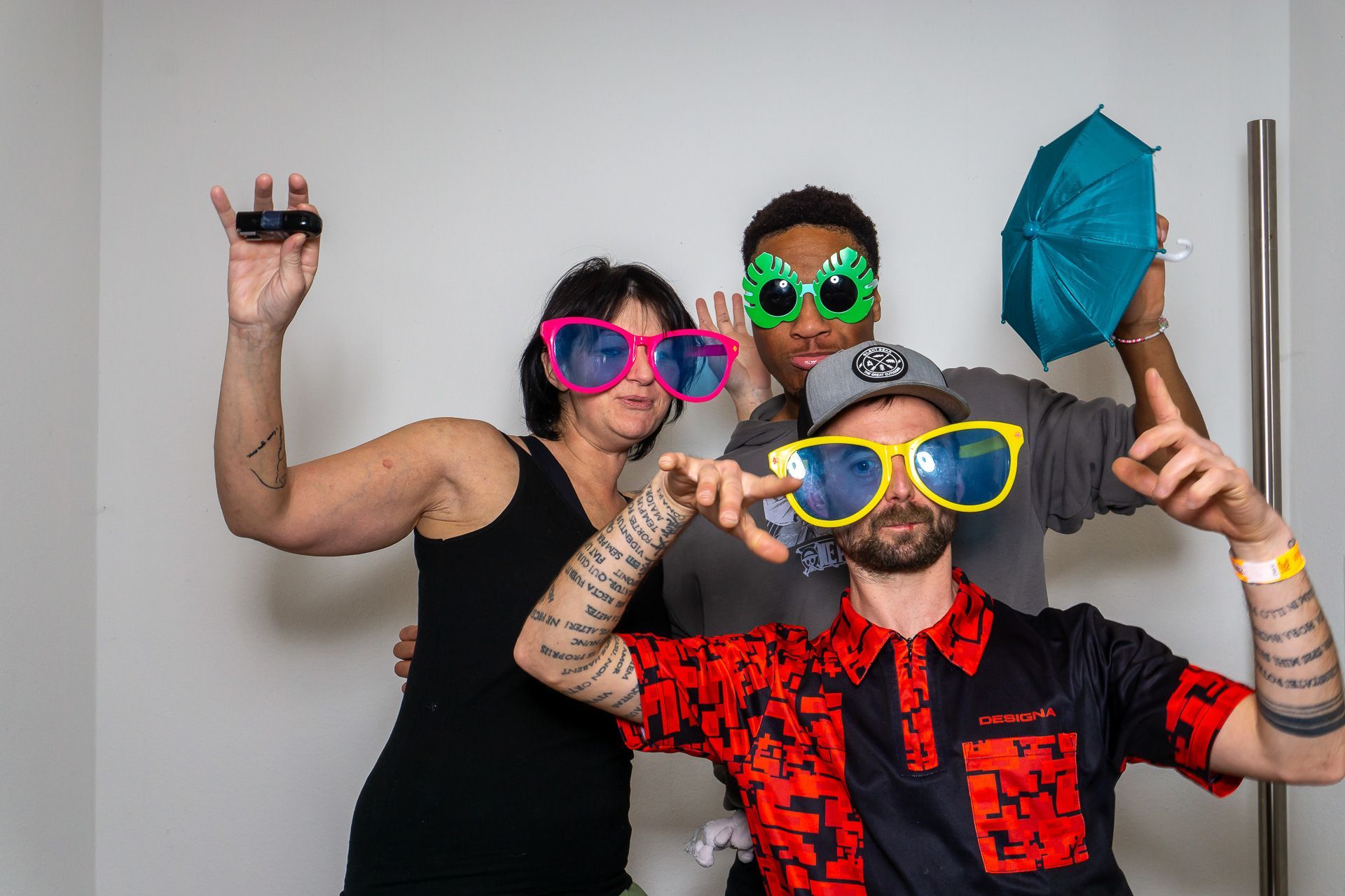 Vier Personen posieren mit Sonnenbrillen und bunten Partyaccessoires und lächeln vor einer schlichten Wand.