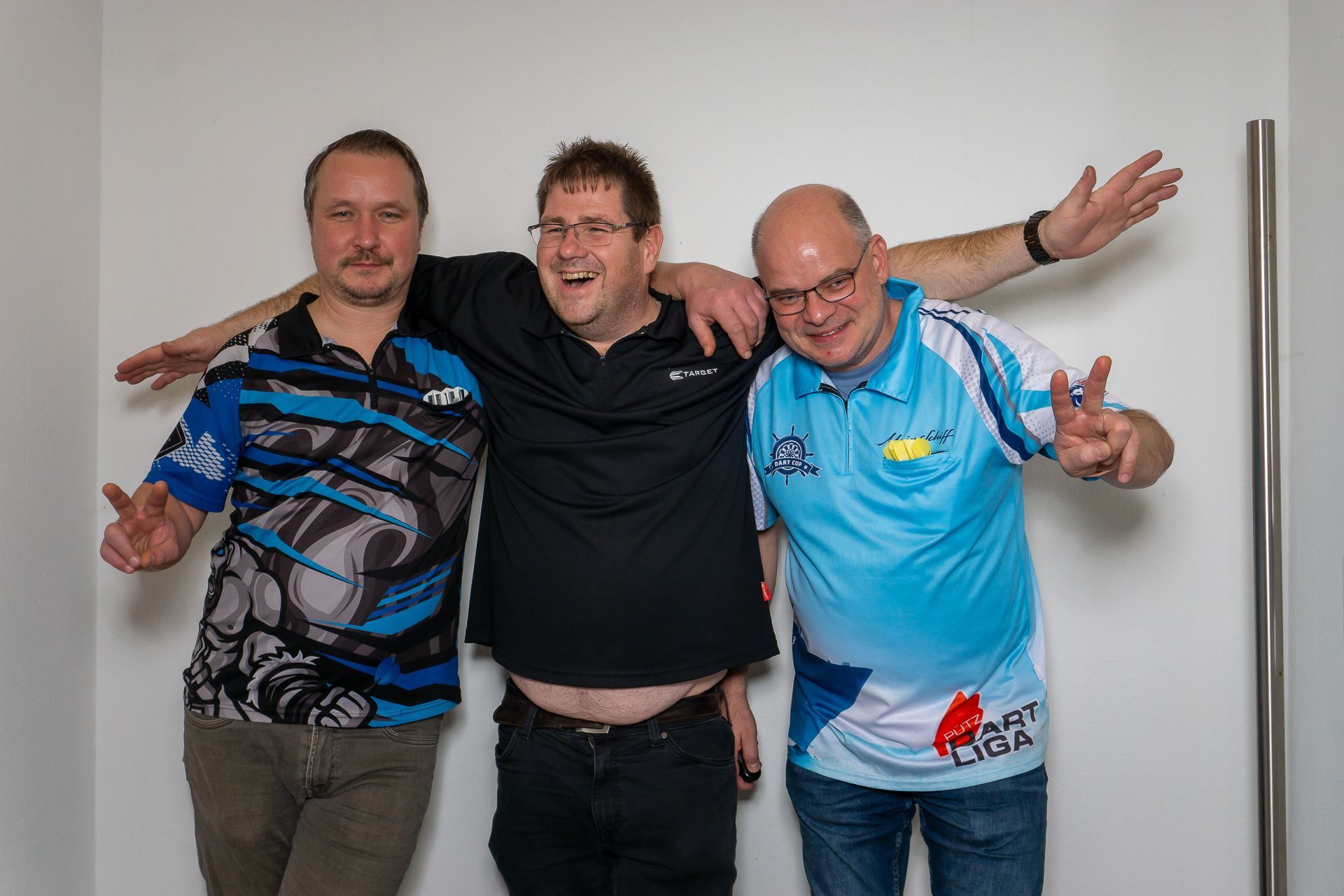Drei Männer posieren mit umeinander geschlungenen Armen vor einer schlichten Wand, lächeln und gestikulieren spielerisch.