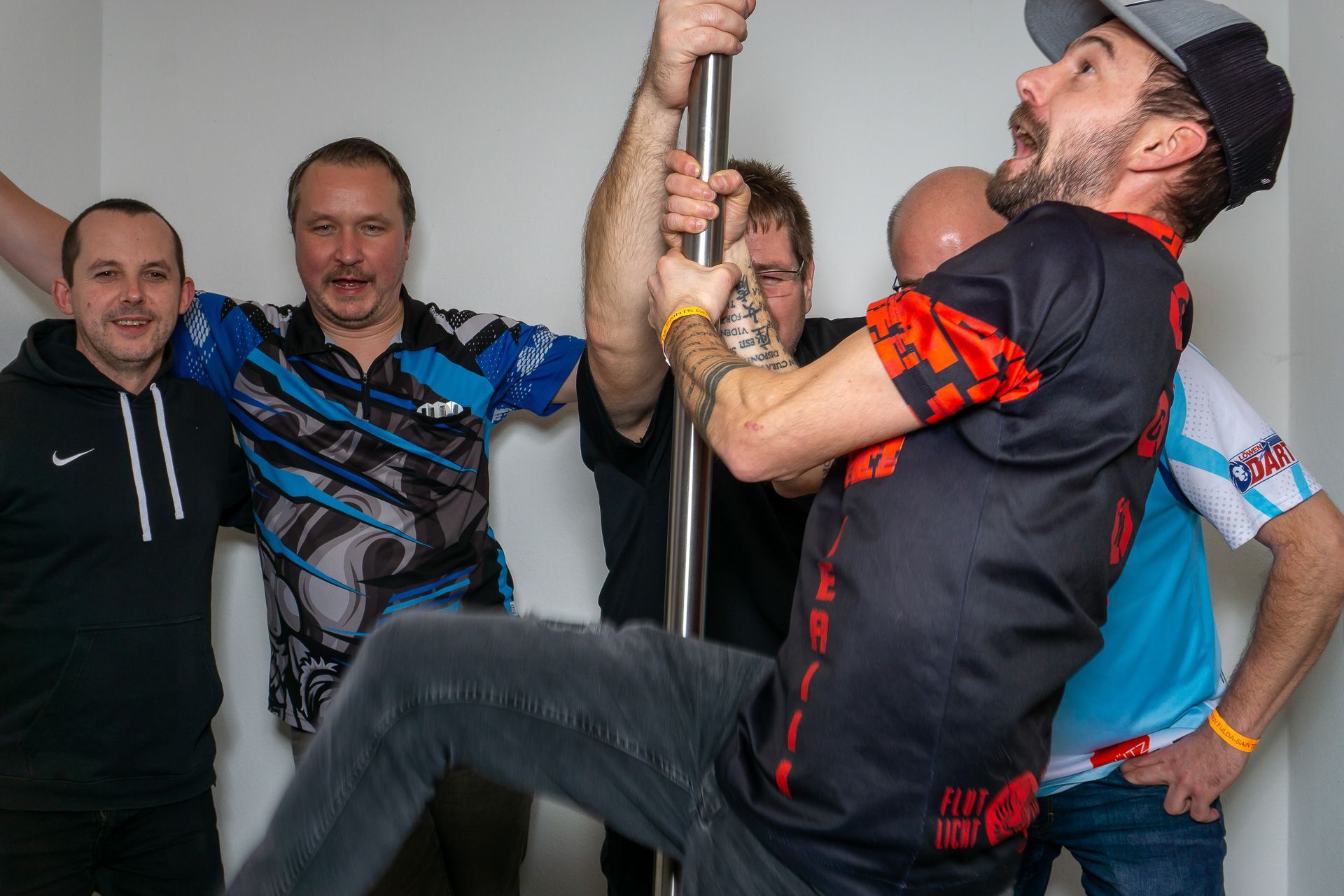 Eine Gruppe von Menschen feiert drinnen; ein Mann hält sich an einer Stange fest und lehnt sich zurück, während die anderen zusehen und lächeln.