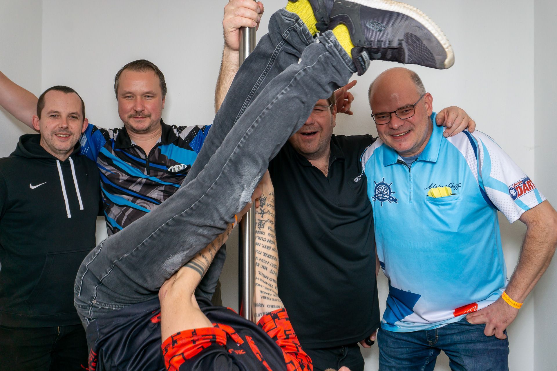 Vier Männer posieren in einem Innenraum, während eine Person in Jeans kopfüber zwischen ihnen gehalten wird.