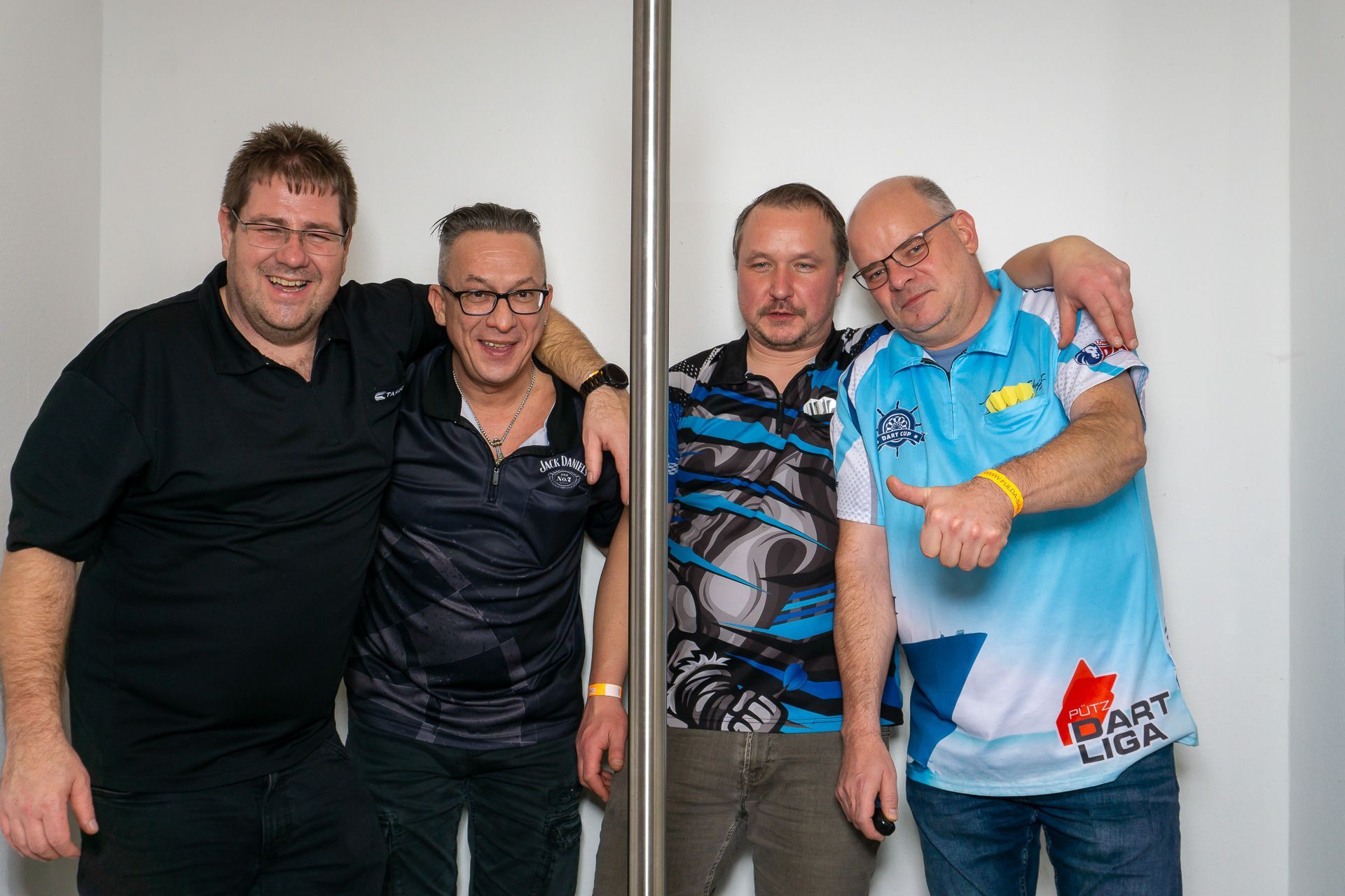 Vier Männer posieren gemeinsam in einem Innenraum, zwei in dunklen Hemden und zwei in blauen Sporttrikots.