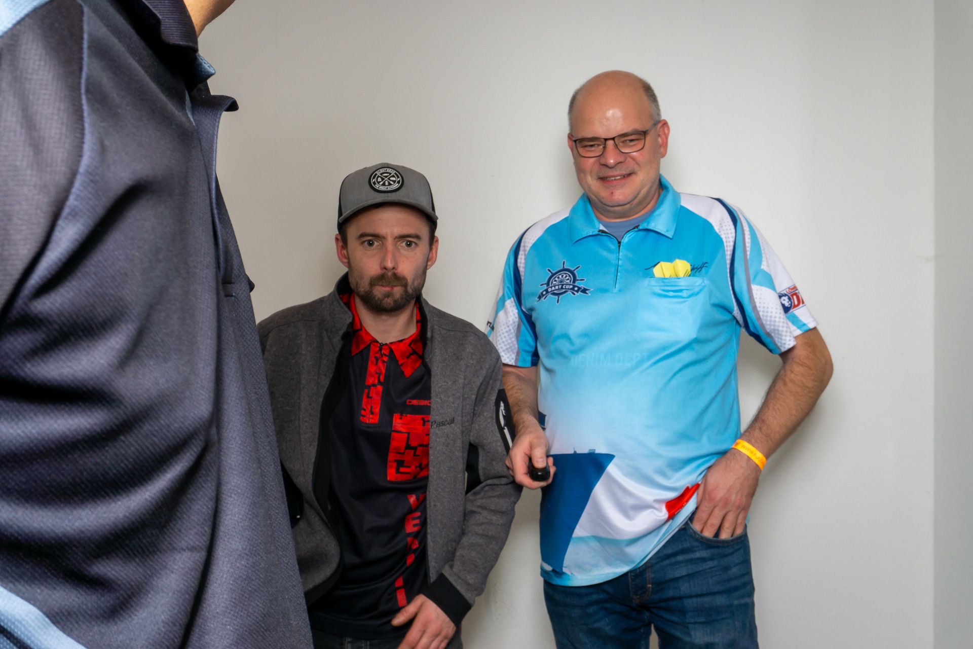 Zwei Männer stehen in einem Innenraum, einer trägt ein blaues Hemd, der andere eine Mütze und eine Jacke, und lächeln in die Kamera.