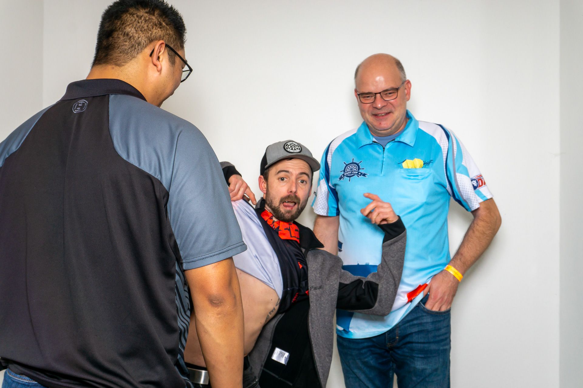 Drei Männer helfen einem sitzenden Mann in einem Haus; einer von ihnen, in einem blauen Hemd, hält seinen Arm.