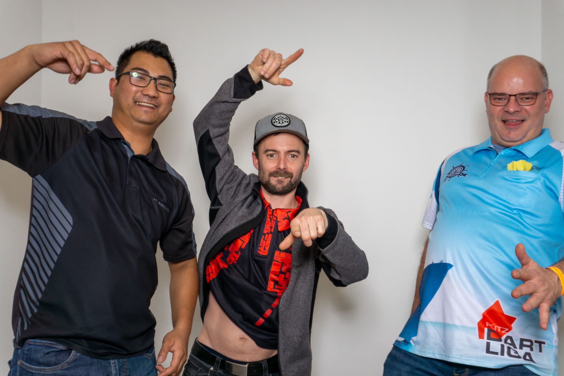 Drei Männer posieren in einem Innenraum, zeigen mit dem Finger und gestikulieren spielerisch; einer trägt ein blaues Trikot, die anderen beiden dunkle Hemden.