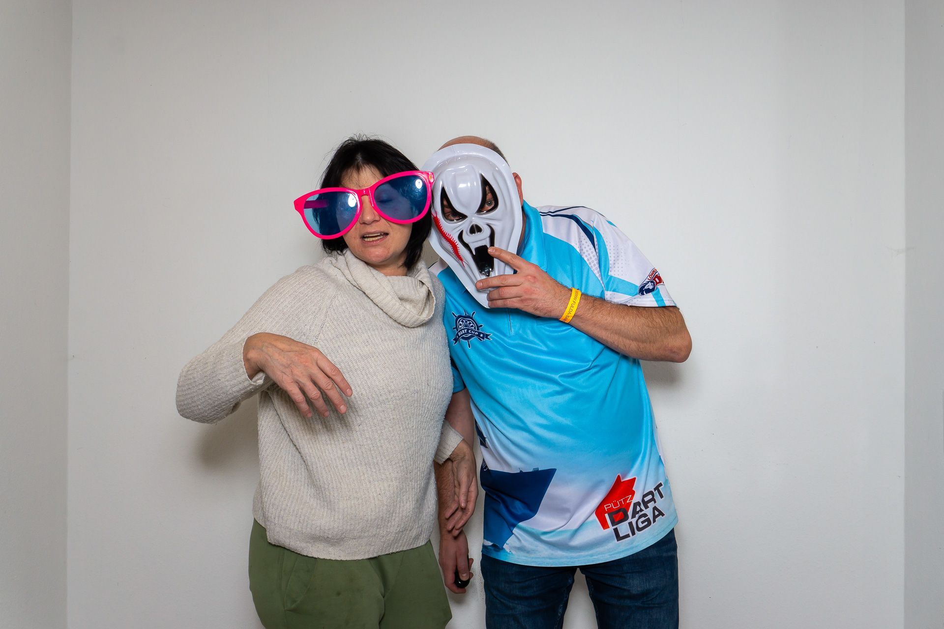 Zwei Personen posieren in einem Innenraum, eine mit einer leuchtend pinkfarbenen Sonnenbrille, die andere mit einer weißen Maske und einem blauen Hemd.
