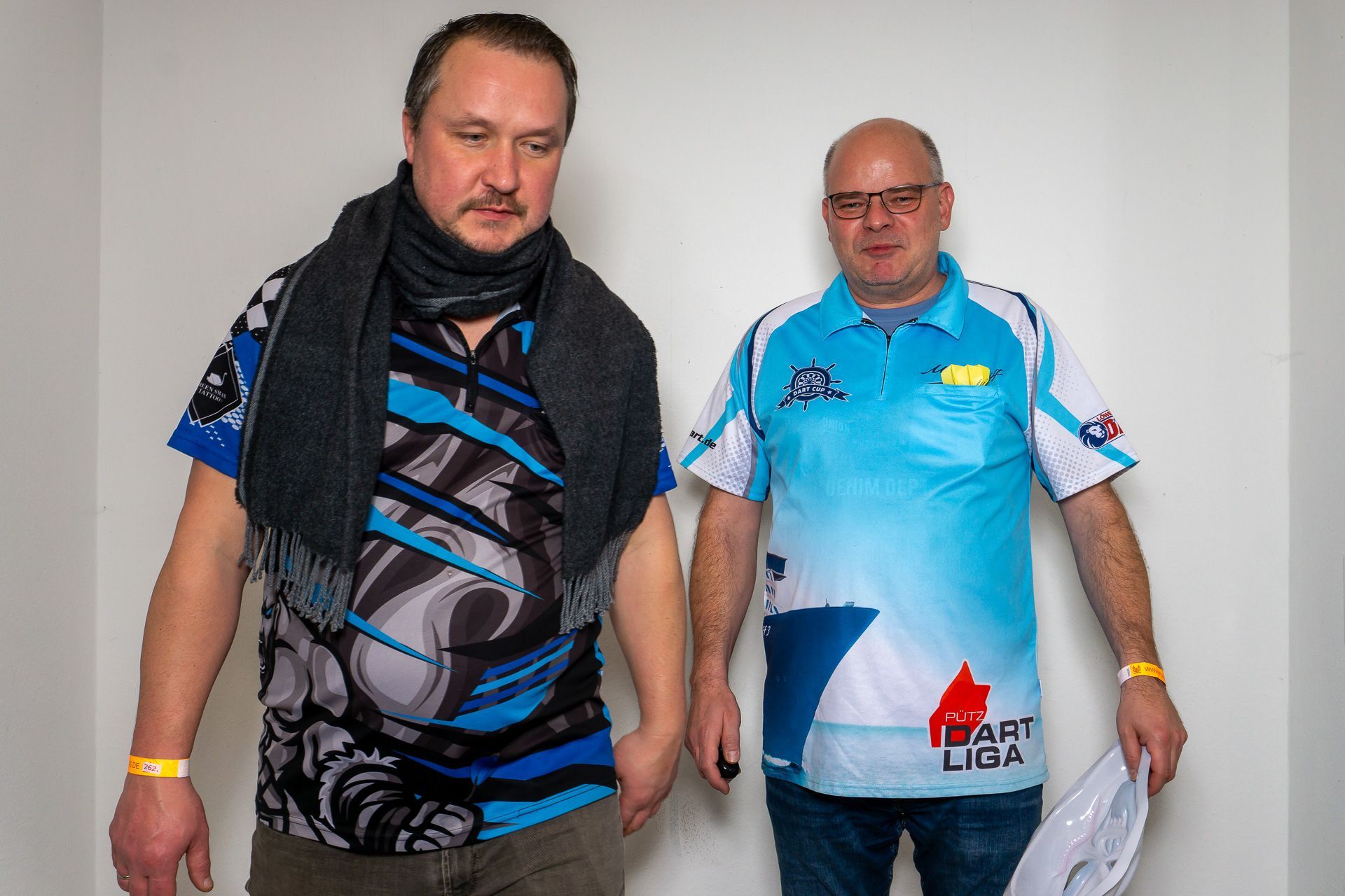 Zwei Männer in blauen und schwarzen Sporttrikots stehen in einem Innenraum und blicken in die Kamera.
