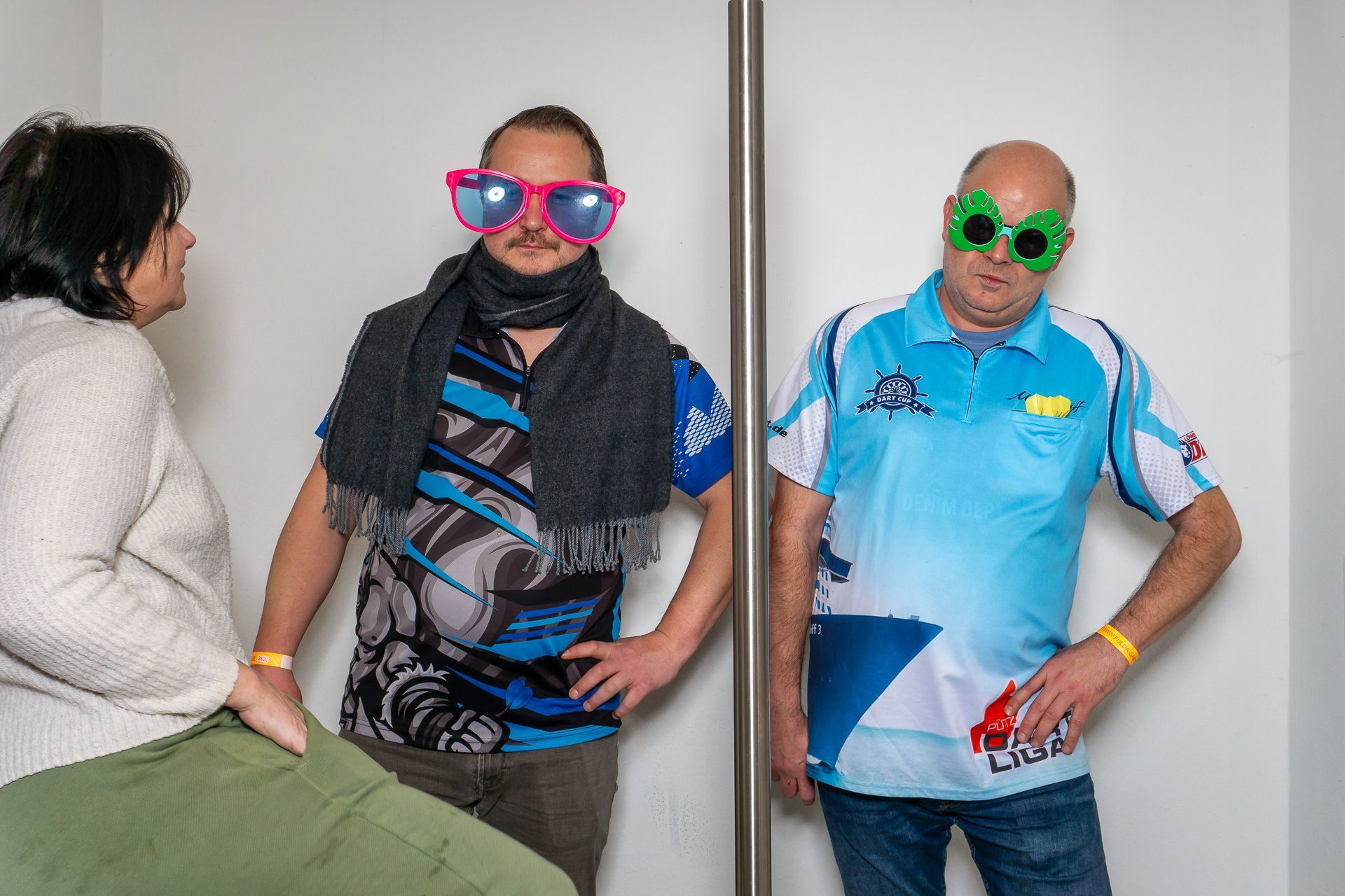 Drei Personen posieren in einem Innenraum, zwei tragen bunte, witzige Brillen und Masken, eine sitzt links.