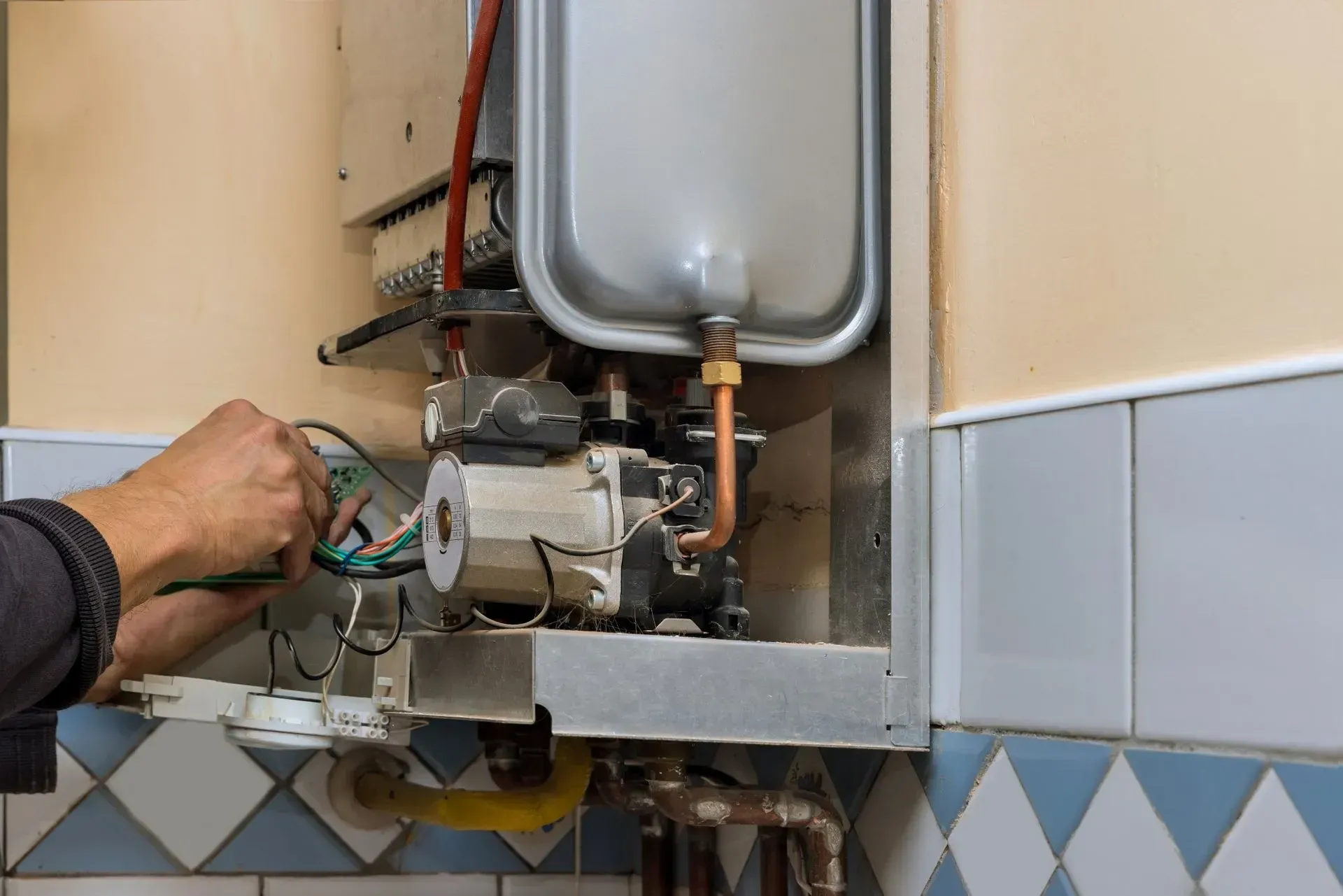 Person repairing a gas water heater, wiring visible, in a utility room.